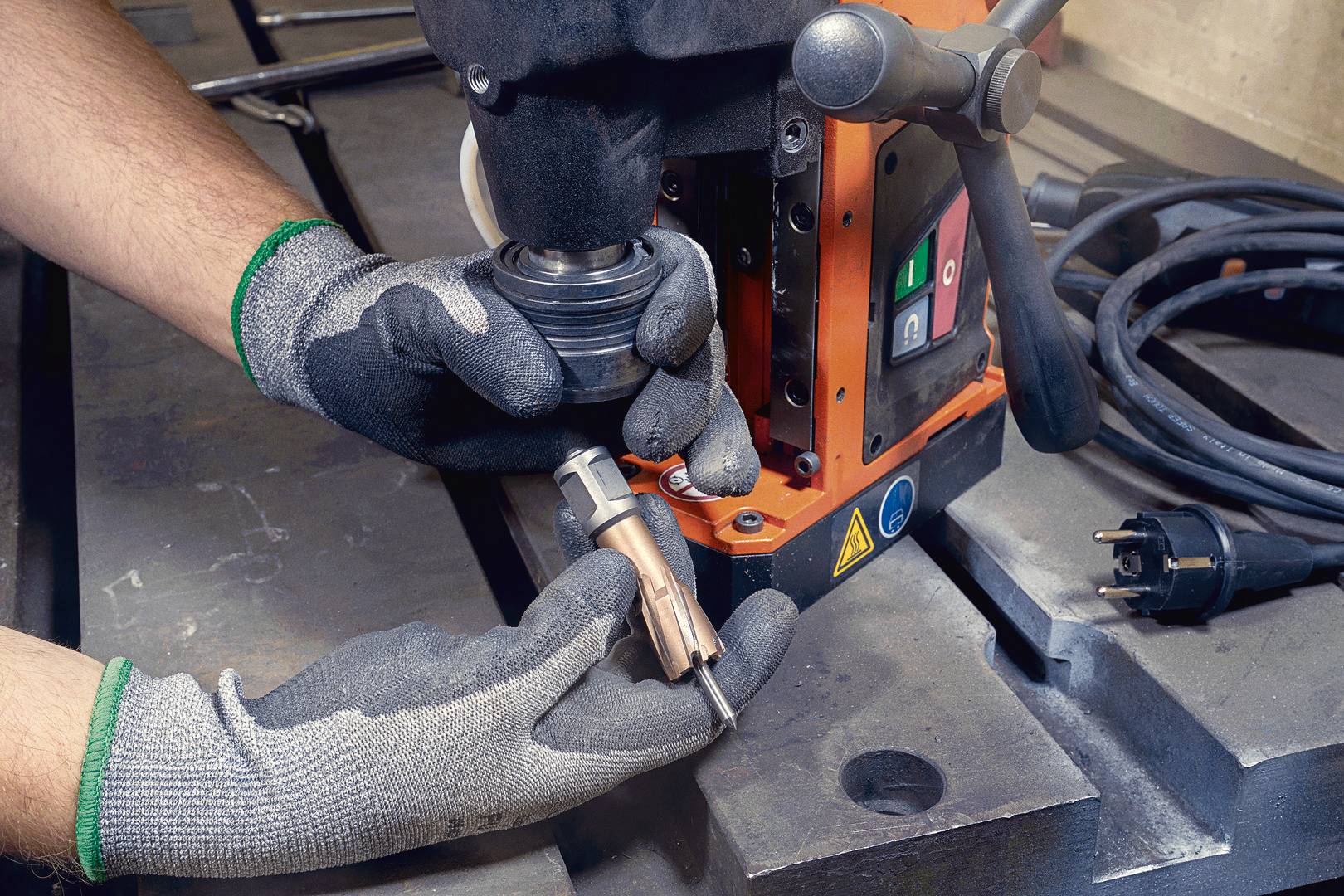 Eine Person trägt Schutzhandschuhe und hält ein Metallteil vor einer industriellen Bohrmaschine, bereit zur Montage.