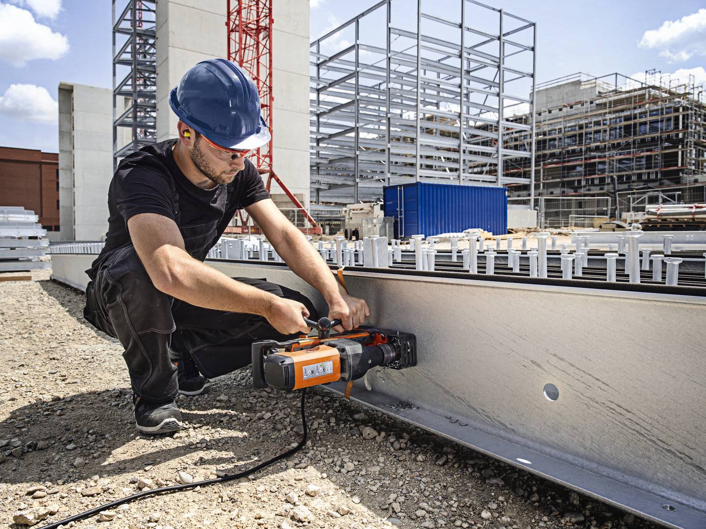 Ein Arbeiter mit blauem Helm montiert mit einem Werkzeug Metallteile auf einer Baustelle, im Hintergrund Gerüste und ein Baukran.