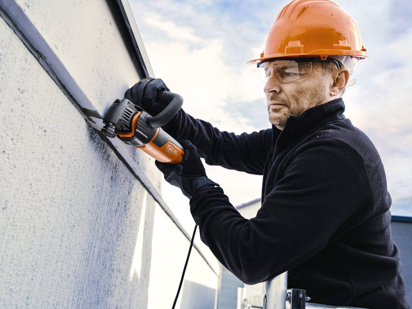 Ein Mann in Sicherheitskleidung arbeitet mit einer Schleifmaschine auf einem Dach. Er trägt einen orangefarbenen Schutzhelm.