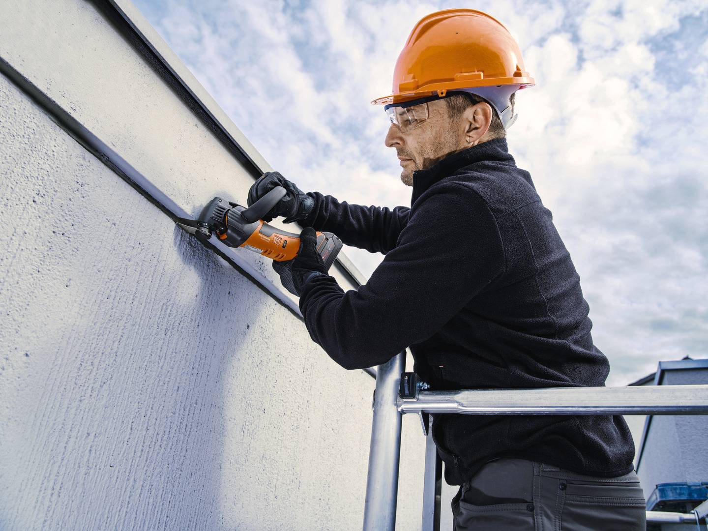 Ein Bauarbeiter mit Schutzhelm und Handschuhen arbeitet mit einem elektrischen Werkzeug an einer Fassade.