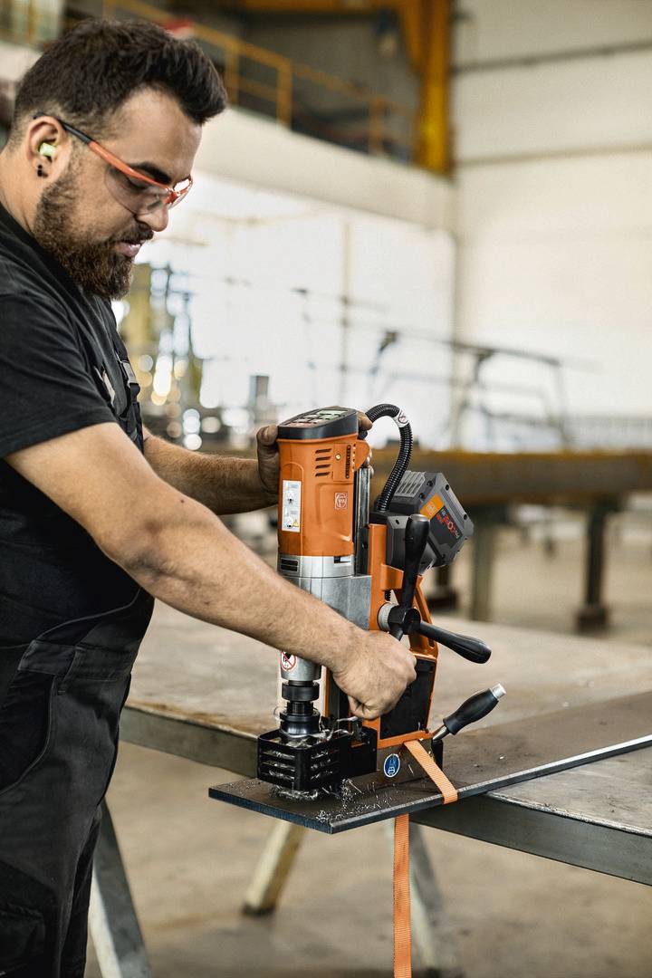 Ein Mann verwendet eine orangefarbene Magnetbohrmaschine, um ein Metallstück auf einer Werkbank in einer Werkstatt zu bearbeiten.
