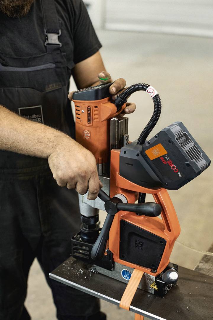 Eine Person bedient eine orange Maschine, die Metall bohrt. Die Szene findet in einer Werkstatt statt.