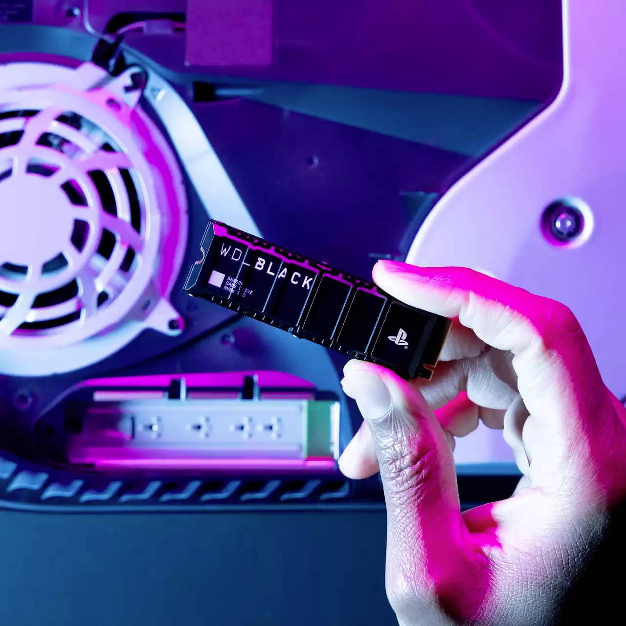 Eine Hand hält einen kleinen schwarzen Speicherchip vor der Rückwand eines geöffneten elektronischen Geräts mit sichtbarem Lüfter.