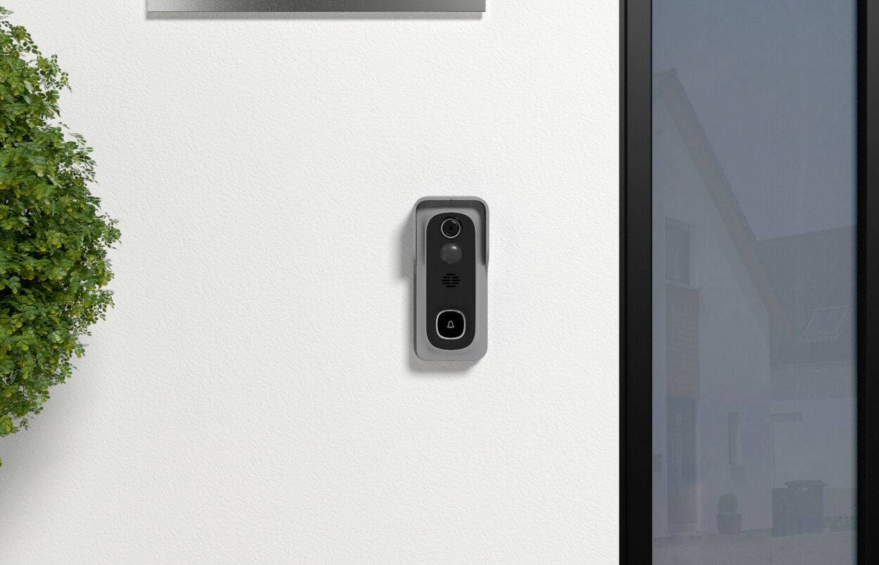 Eine Außenwand mit einer modernen Video-Türklingel, die neben einem Fenster montiert ist. Links ist ein Teil einer grünen Pflanze sichtbar.