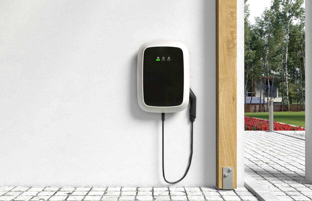 Ladestation für Elektroautos an einer weißen Wand montiert, mit grünem Licht. Kabel hängt herab. Garten im Hintergrund sichtbar.