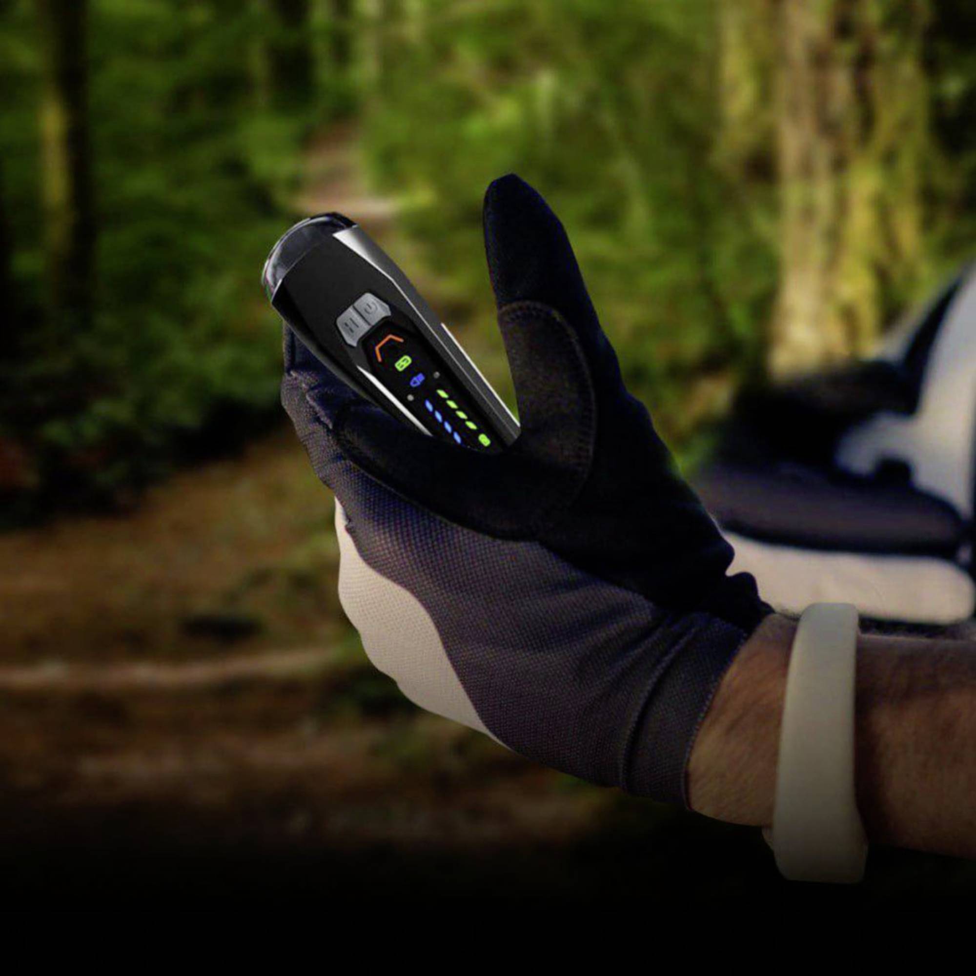 Eine behandschuhte Hand hält ein tragbares Gerät mit mehreren leuchtenden Anzeigen. Hintergrund unscharf, vermutlich Waldweg.