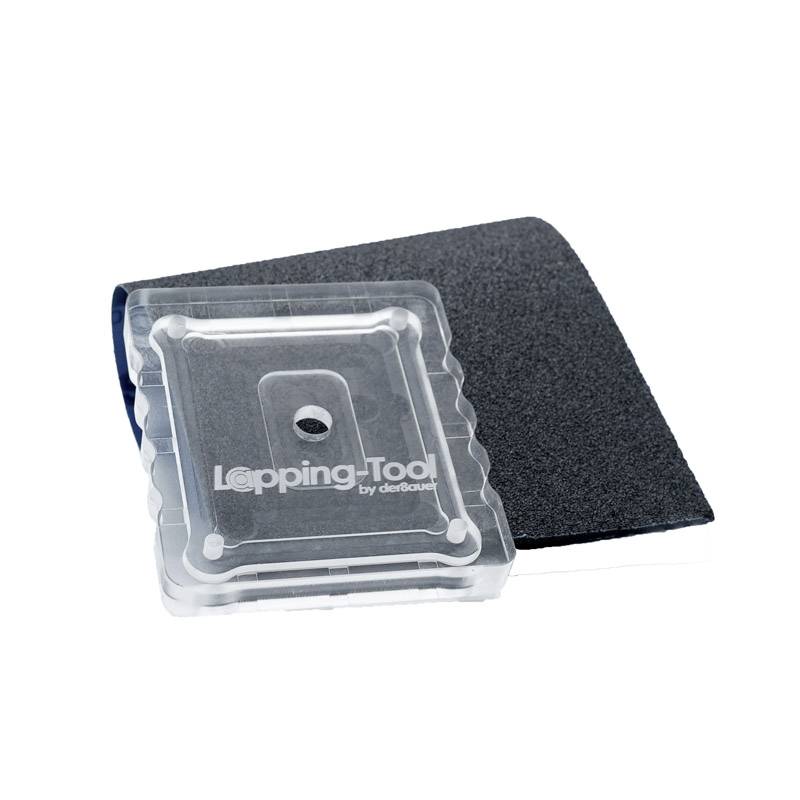 Ein quadratisches Schleifwerkzeug mit transparentem Gehäuse liegt auf einem schwarzen Schleifpad. Text auf dem Werkzeug: 'Lapping-Tool'.