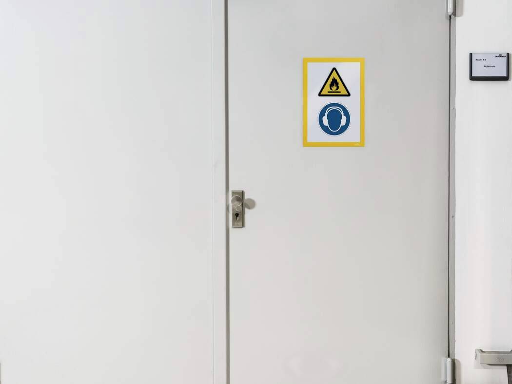 Weiße Tür mit Sicherheitszeichen: 'Warnung vor Lärmbelästigung' und 'Gehörschutz benutzen'. Das Schild ist deutlich sichtbar.