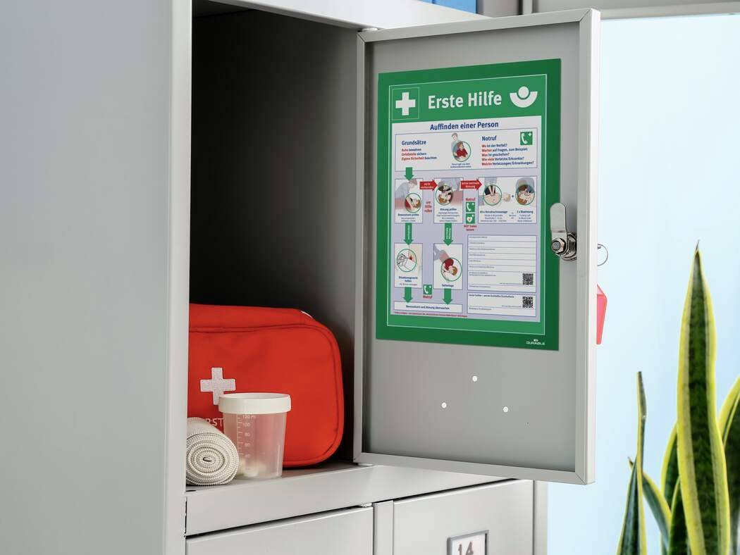 Ein geöffneter Erste-Hilfe-Schrank zeigt ein Erste-Hilfe-Set mit Verband und Becher. An der Tür hängt ein Plakat mit Anweisungen.