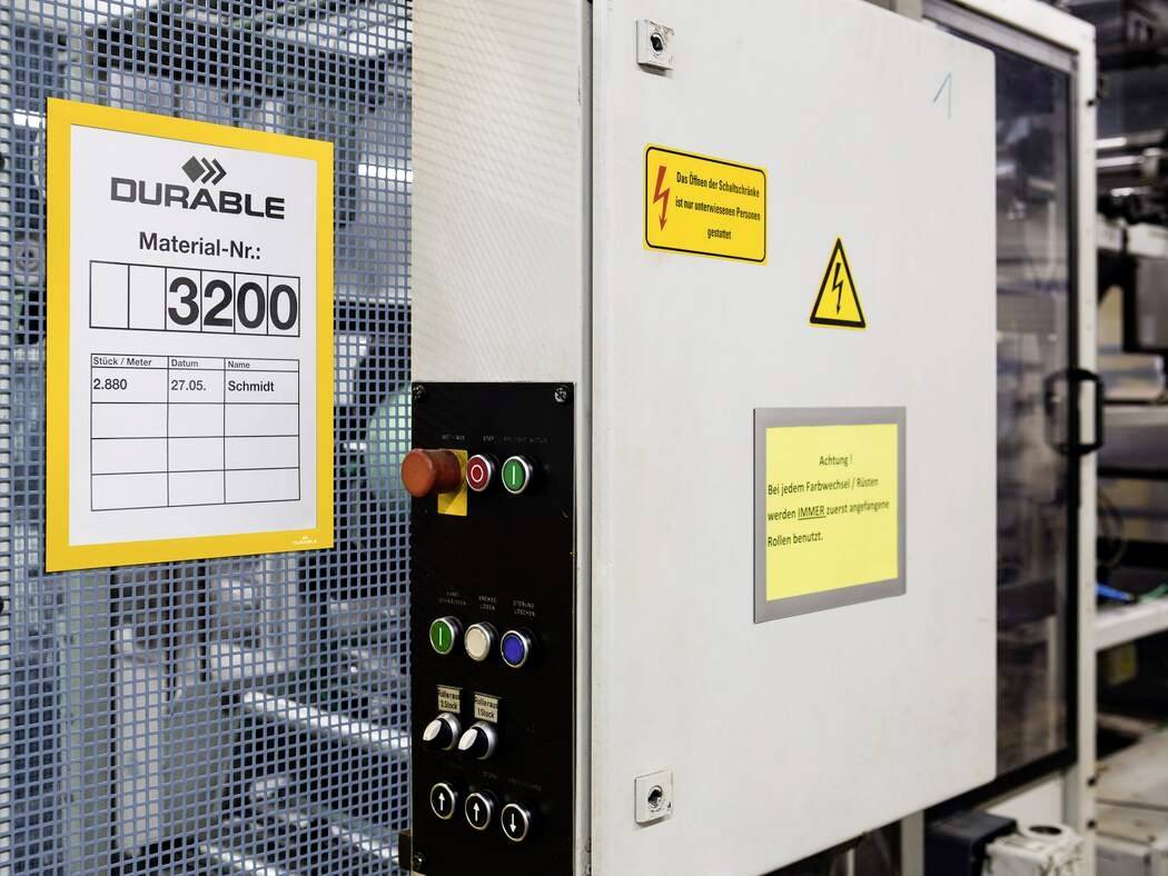 Maschinensteuerung mit Notfallknöpfen und Anzeigetafel mit Materialnummer '3200'. Wichtige Schilder: Warnung vor Elektrizität.