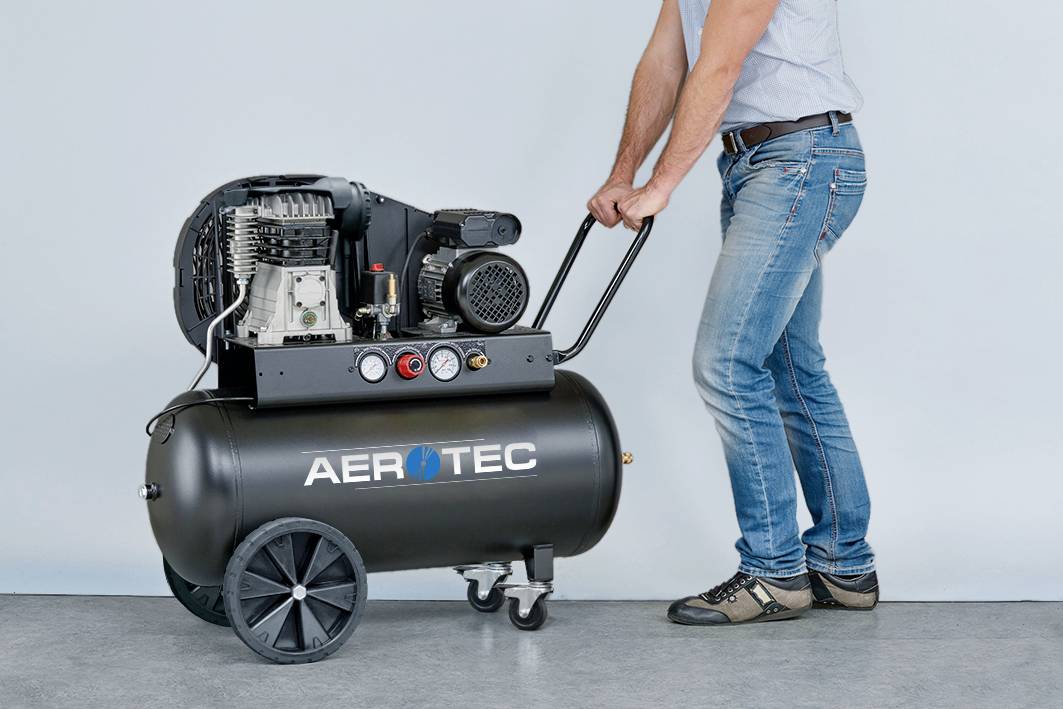 Ein Mann in Jeans schiebt einen schwarzen Luftkompressor mit 'AEROTEC'-Schriftzug auf zwei Rädern über einen grauen Boden.