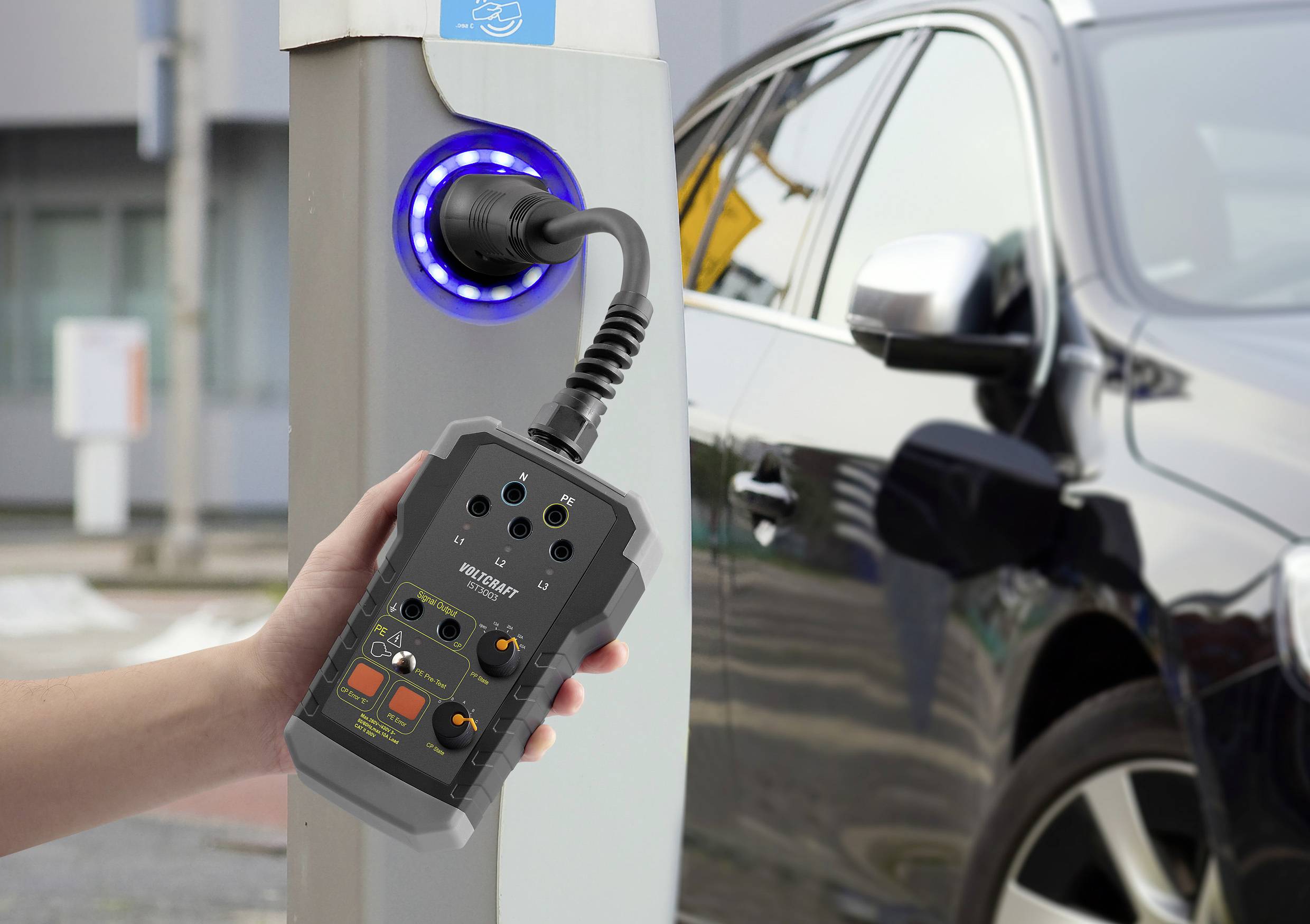 Eine Hand hält ein Messgerät, das an eine Ladestation angeschlossen ist, während ein Auto geladen wird.