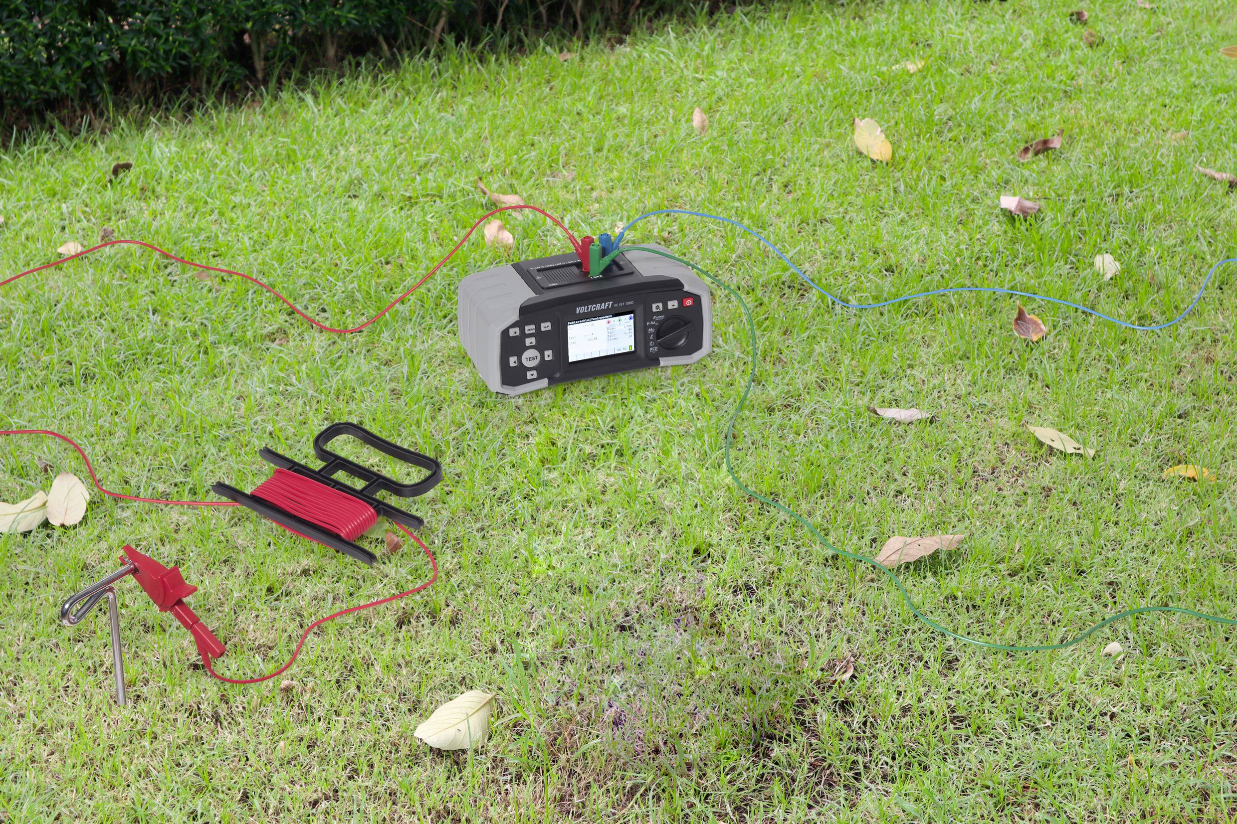 Ein digitales Erdmessgerät liegt auf grünem Gras, verbunden mit mehreren Kabeln und einem Erdungsspieß. Blätter sind auf dem Boden verstreut.