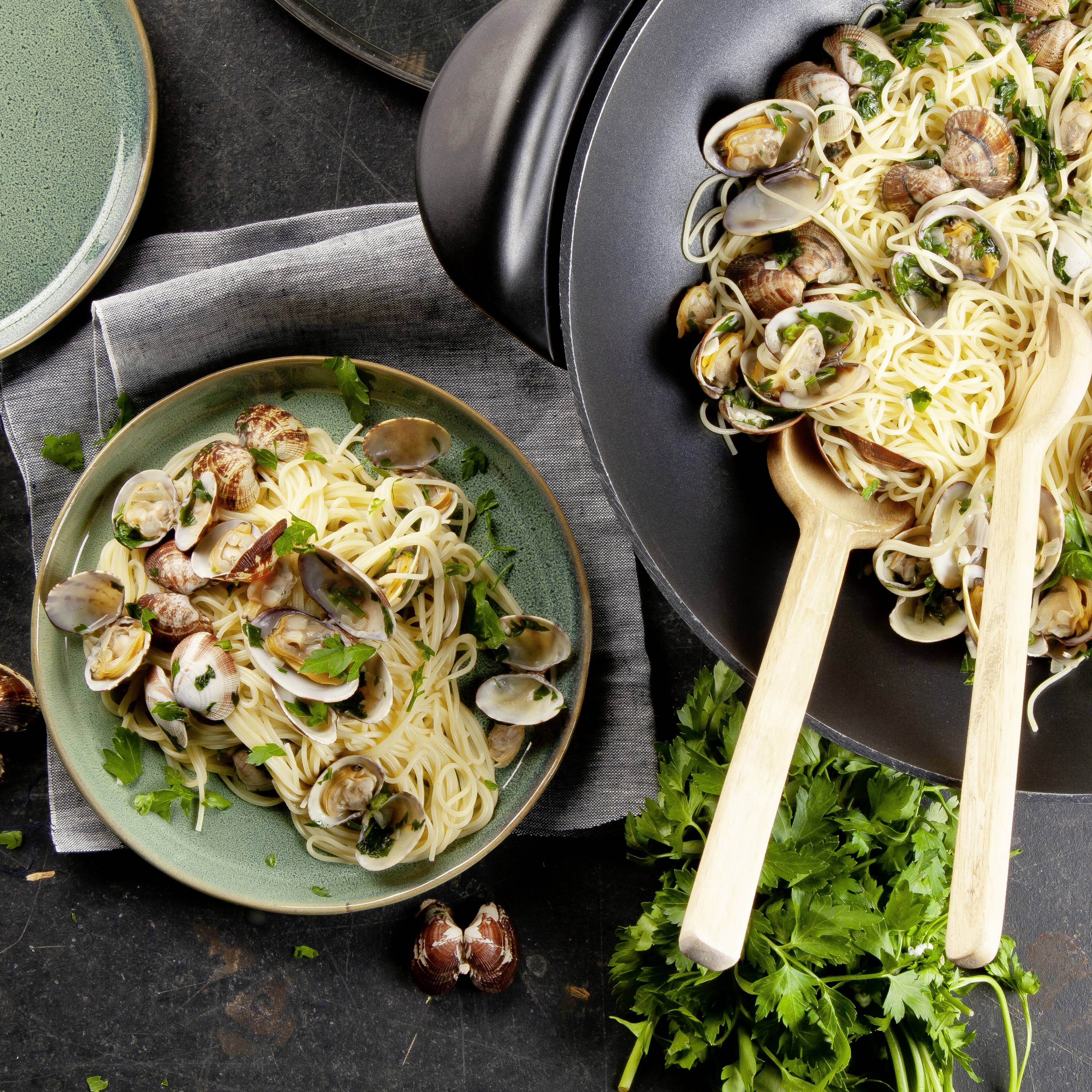Nudeln mit Muscheln, Petersilie garniert, in schwarzer Pfanne serviert; daneben gefüllter Teller und frische Petersilie auf dunklem Tisch.