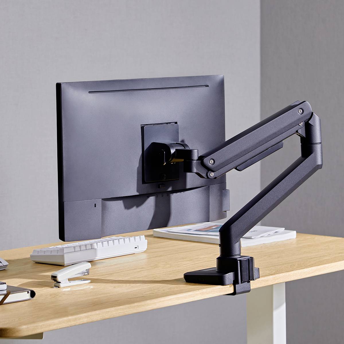 Ein Computermonitor mit einem flexiblen, ergonomischen Monitorarm ist auf einem Schreibtisch montiert, der auch eine Tastatur und Bücher enthält.