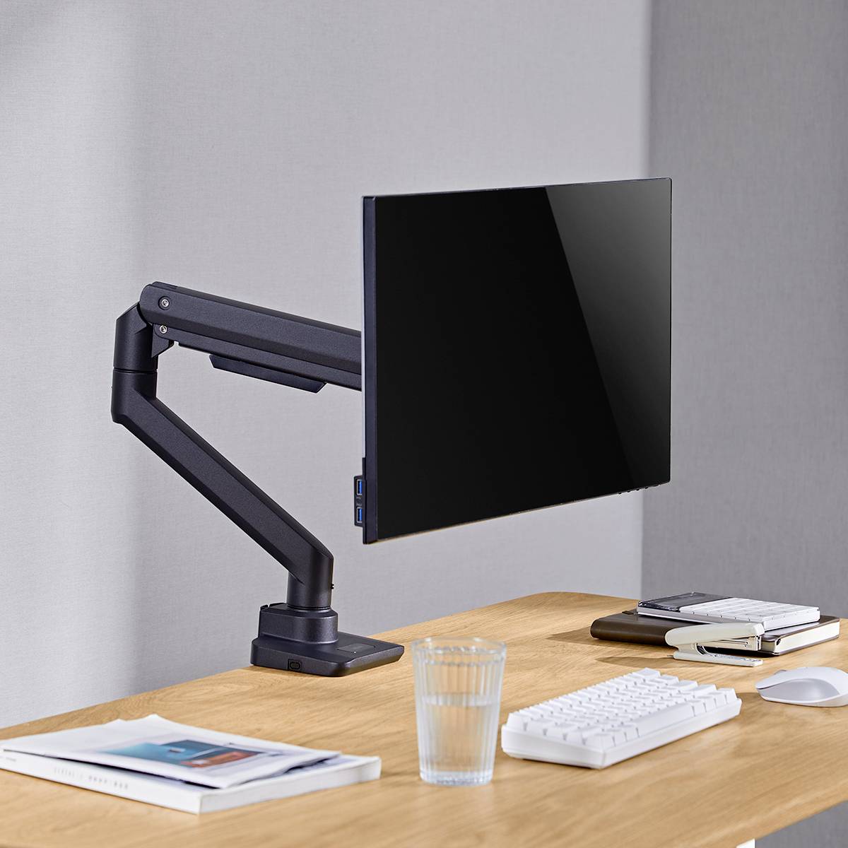 Ein moderner Schreibtisch mit einem Computerbildschirm an einem Gelenkarm, einer Tastatur, einer Maus, einem Glas Wasser und Notizbüchern.