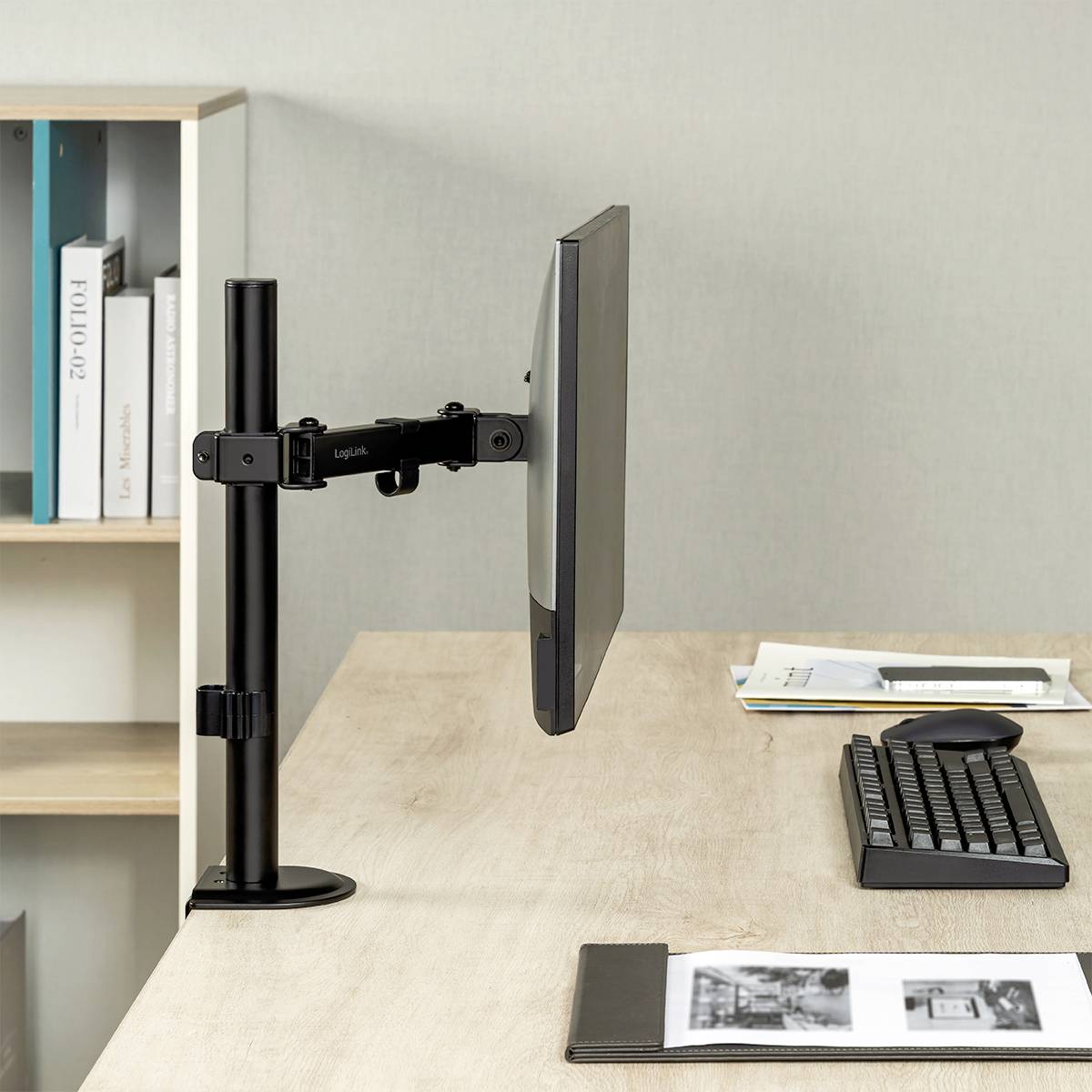 Ein Computerbildschirm auf einem verstellbaren Arm steht auf einem Schreibtisch neben einer Tastatur. Im Hintergrund ist ein Bücherregal.