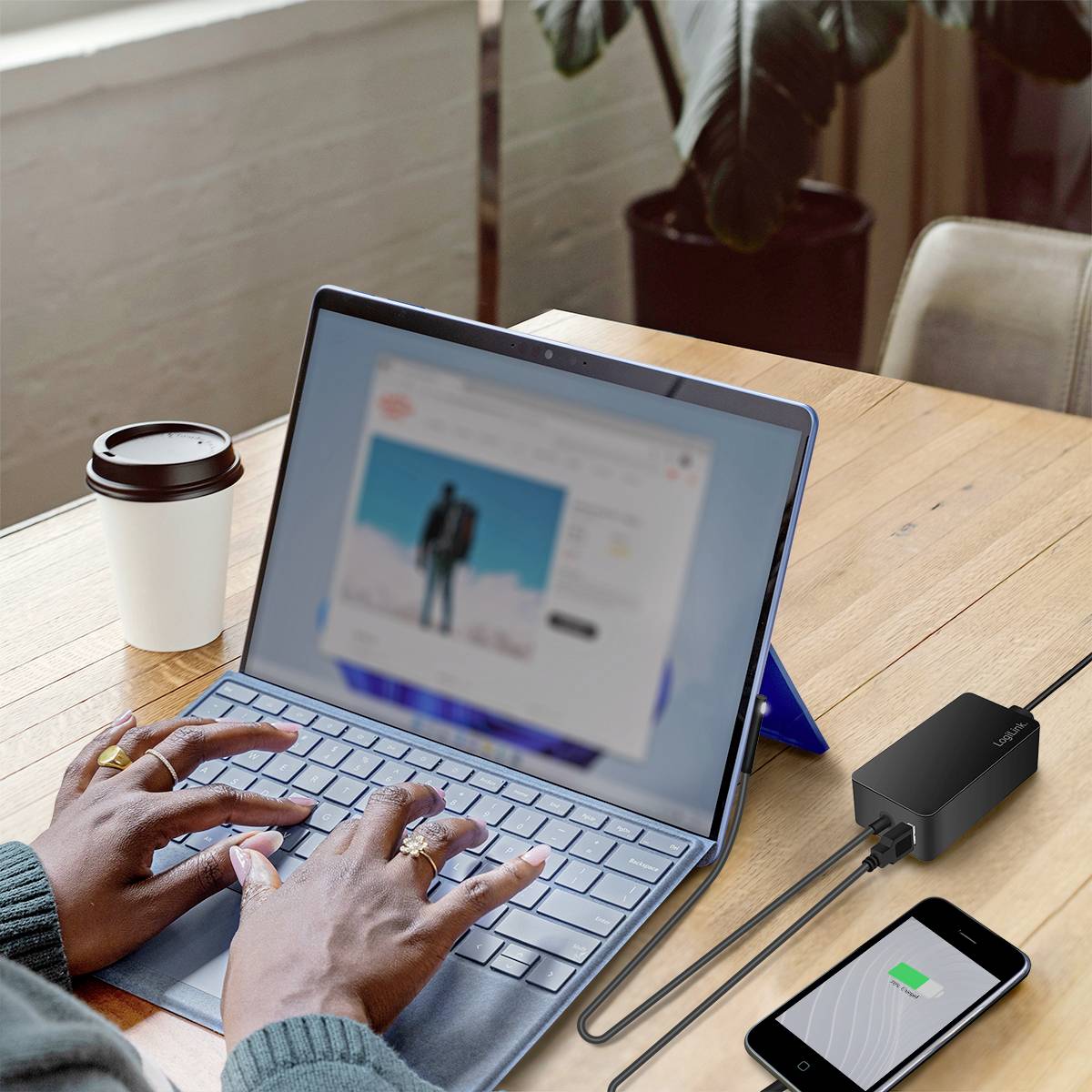 Person arbeitet an einem Laptop mit Online-Shop; daneben ein Smartphone mit Ladeanzeige und eine Tasse Kaffee auf dem Tisch.