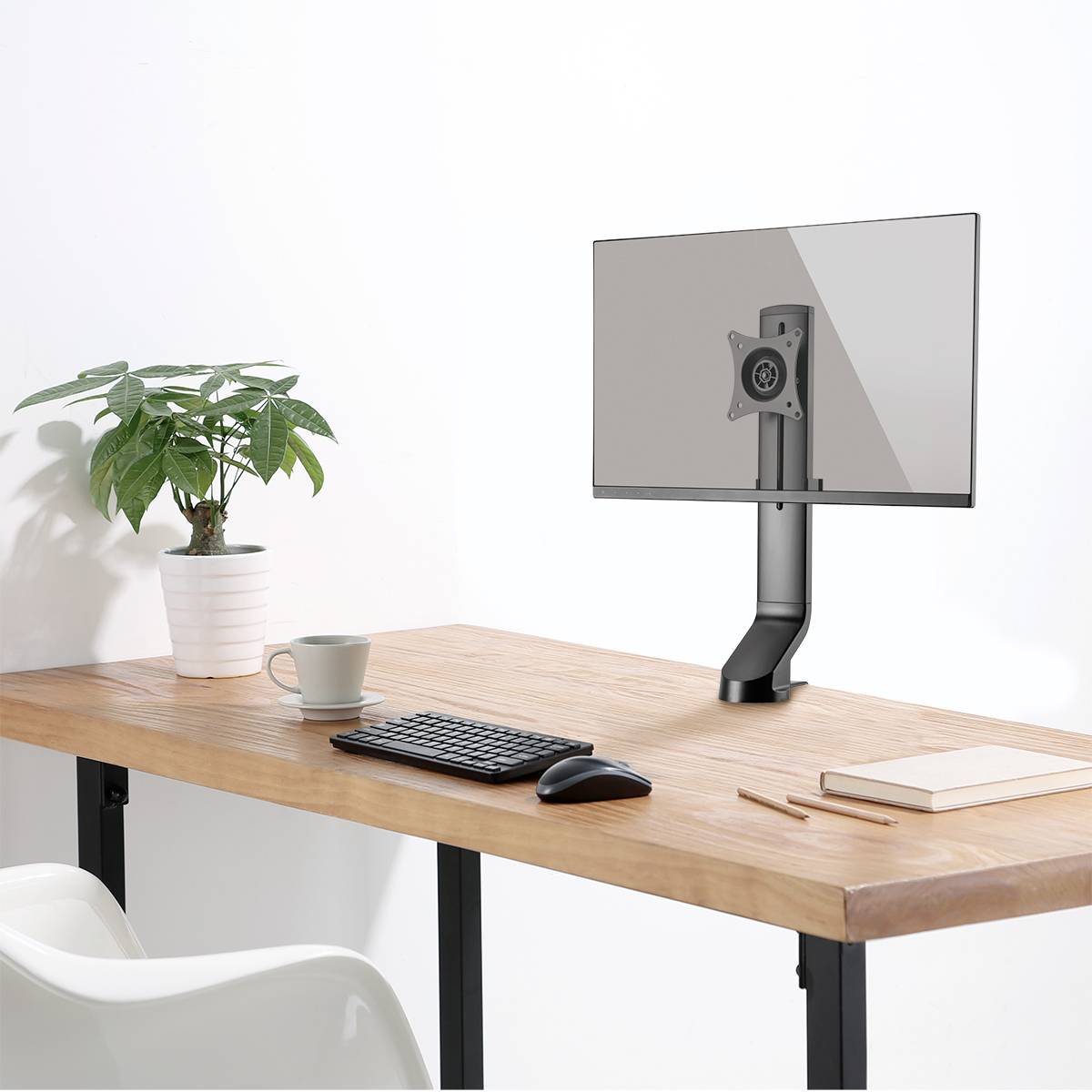Auf einem Schreibtisch steht ein Monitorhalter ohne Bildschirm. Daneben eine Tastatur, eine Maus, ein Buch und eine Pflanze in einem Topf.