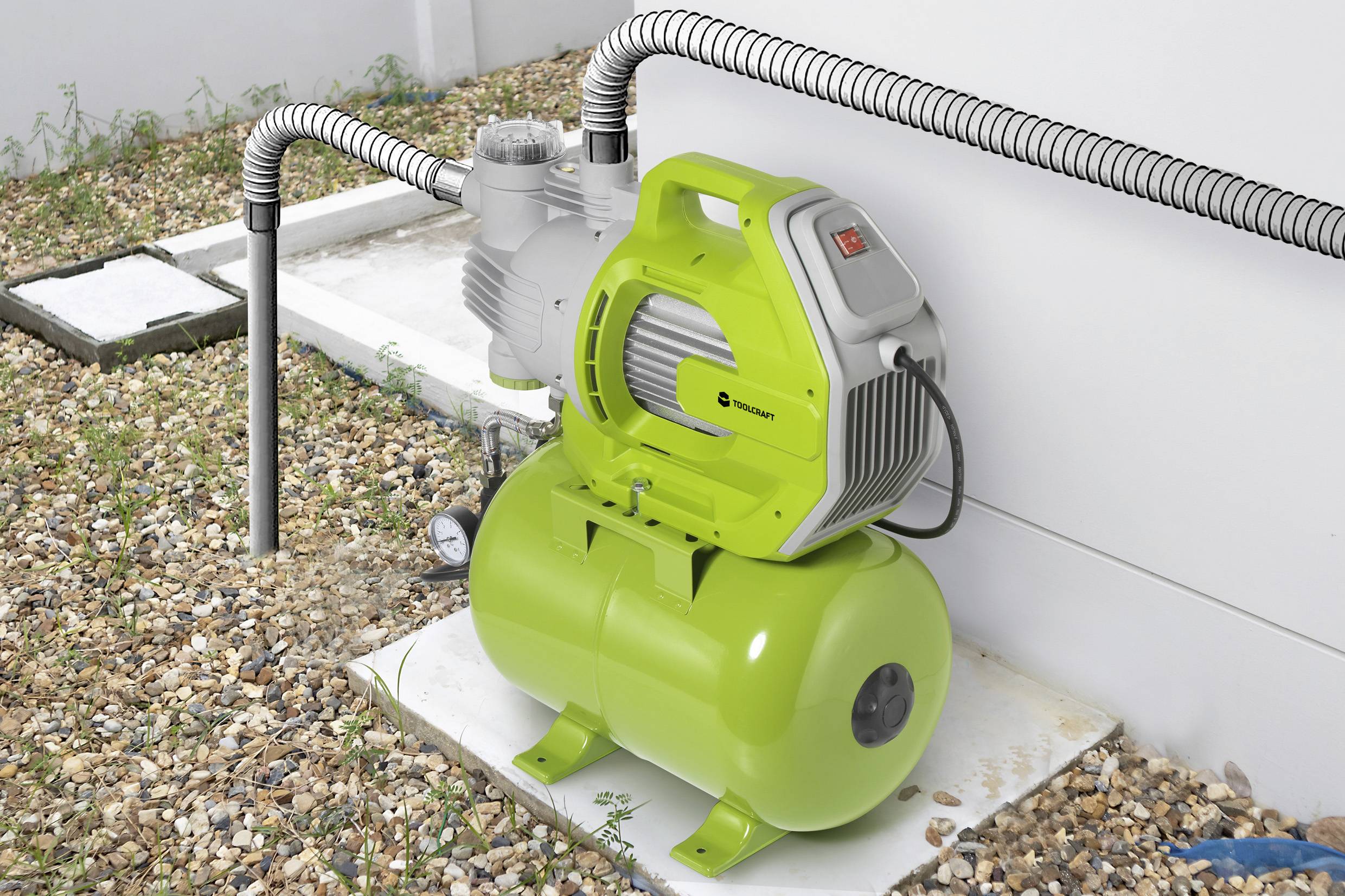 Eine grüne Wasserpumpe mit einem Tank ist an Rohre angeschlossen. Sie steht auf einem Kiesboden neben einer weißen Wand.