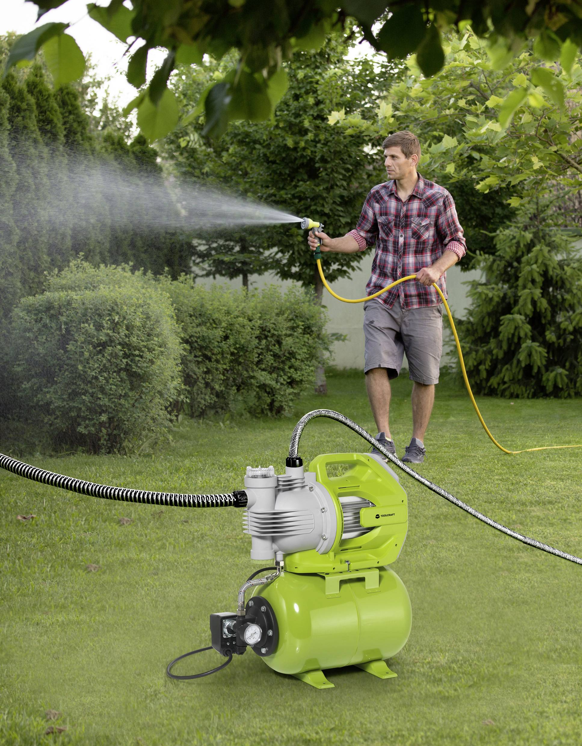 Ein Mann bewässert einen Rasen mit einem Wasserschlauch, während eine grüne Wasserpumpe im Vordergrund zu sehen ist.