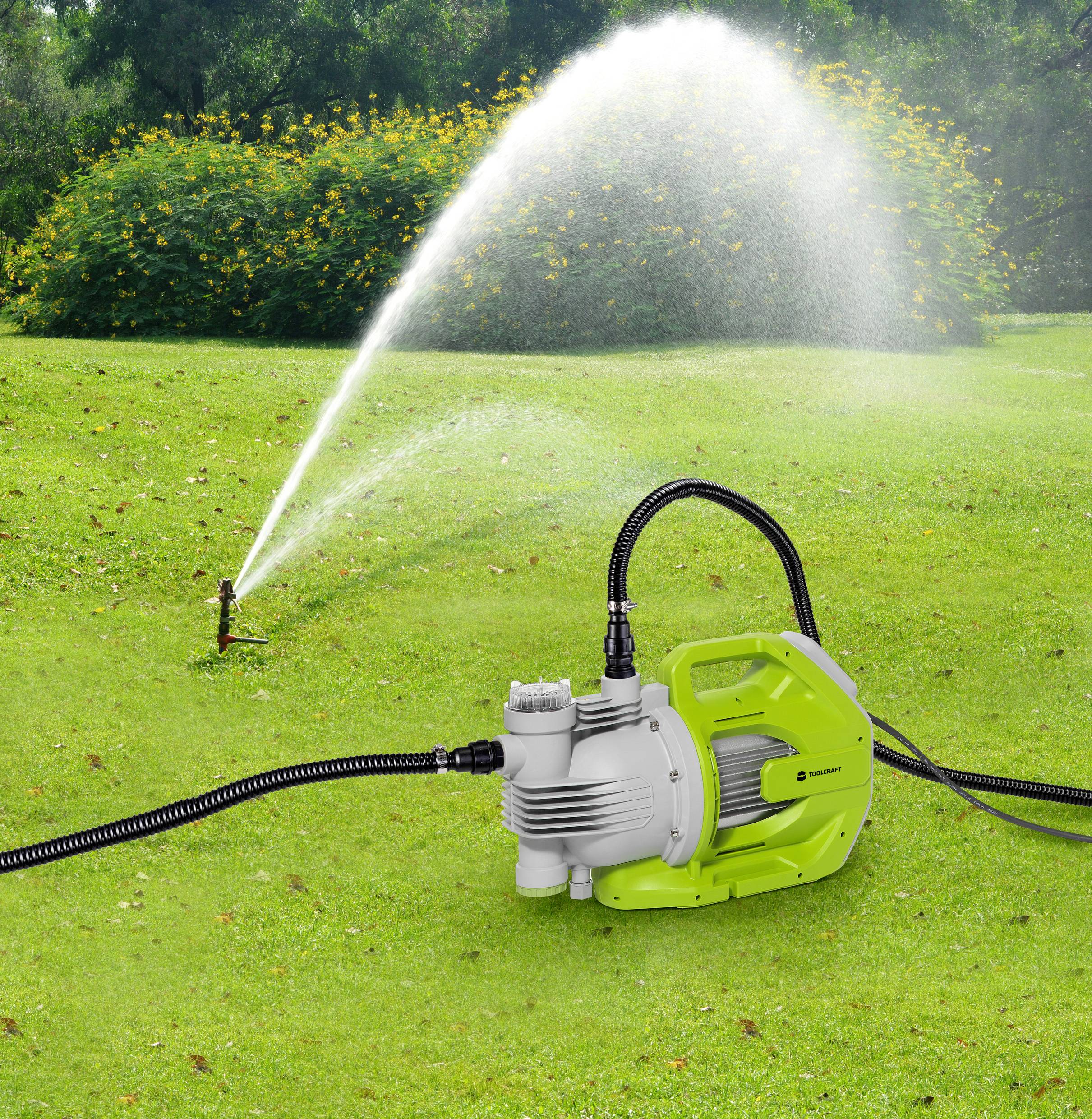 Grüne Wasserpumpe auf Rasen, verbunden mit Schlauch, der Wasser zu einem Sprinkler leitet, um den Garten gleichmäßig zu bewässern.