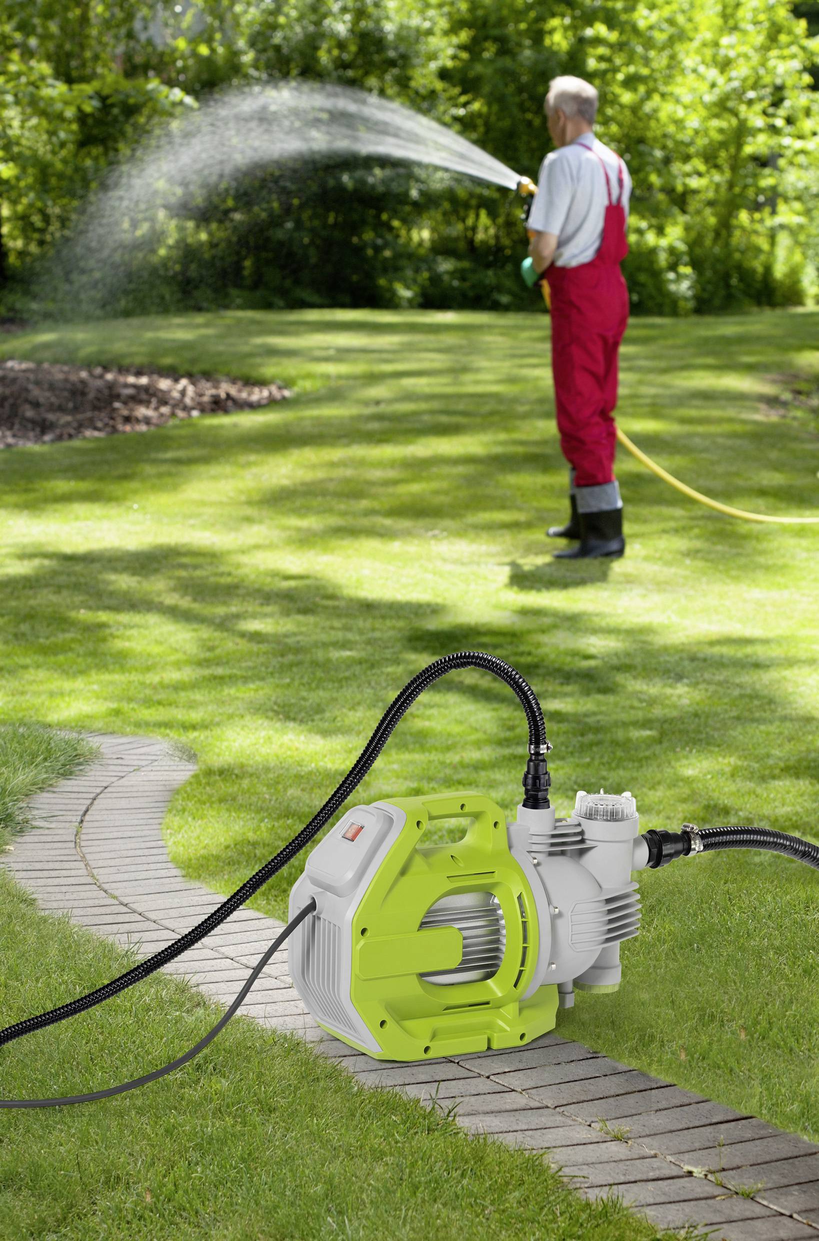 Mann in rotem Overall bewässert Rasen mit Gartenschlauch. Im Vordergrund steht eine grüne elektrische Wasserpumpe auf einem Weg.