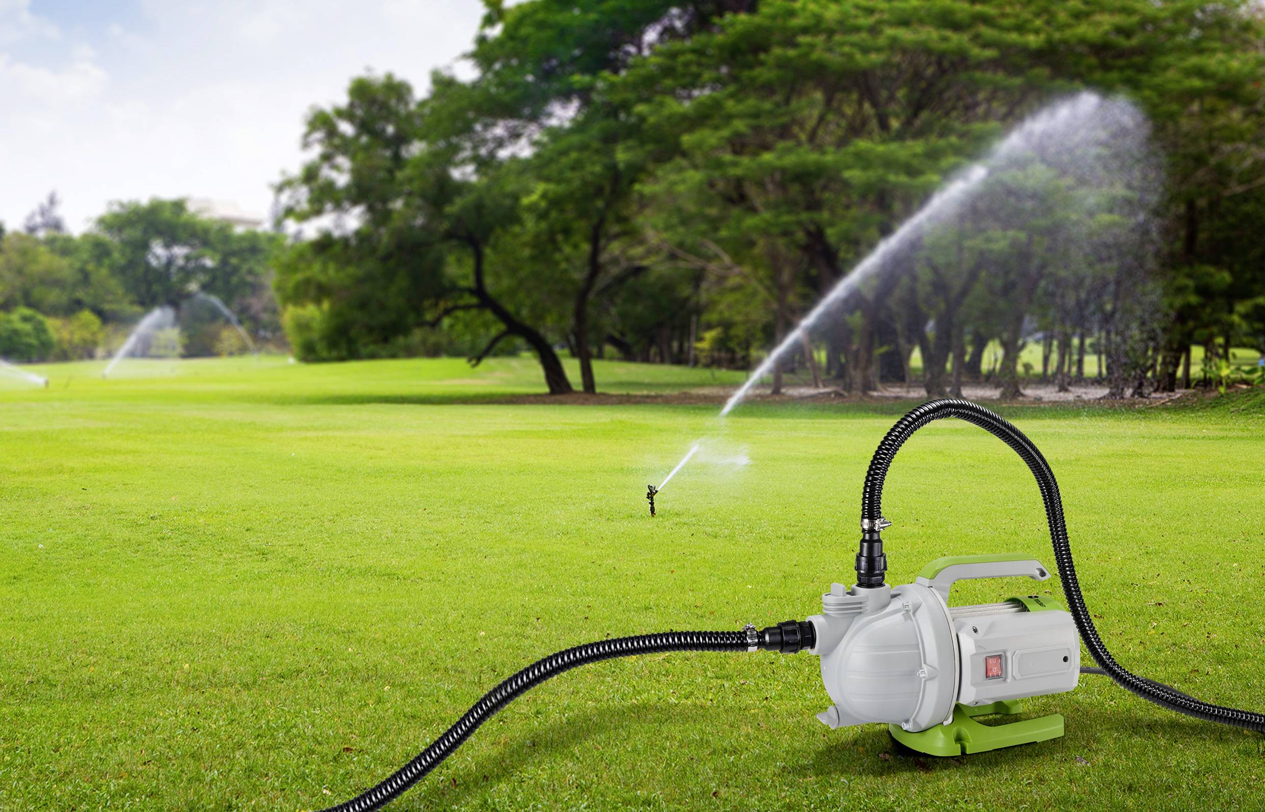 Eine Rasenpumpe mit Schläuchen im Vordergrund bewässert eine große, grüne Rasenfläche mit mehreren Sprinkleranlagen im Hintergrund.