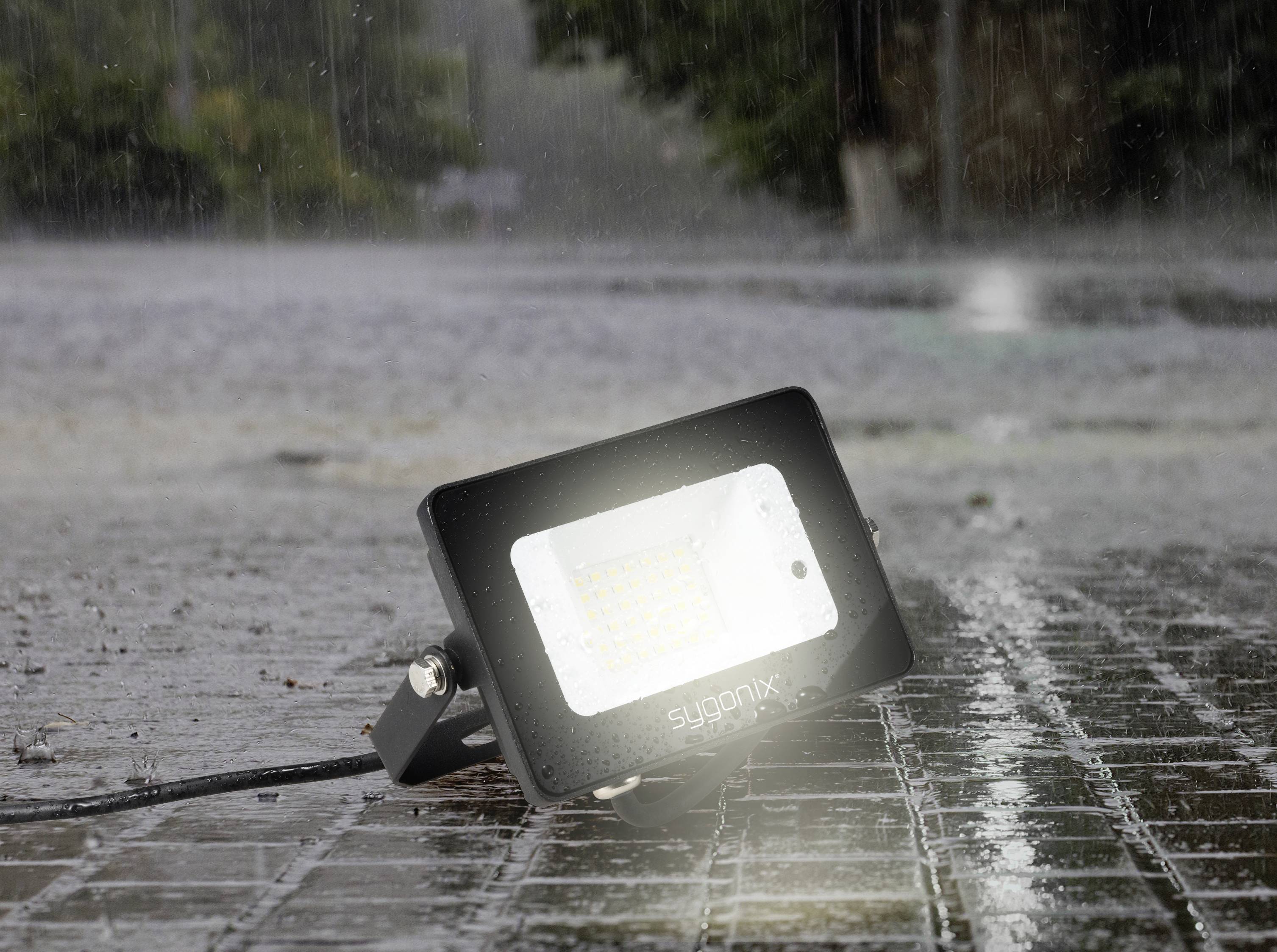 Ein leuchtender LED-Strahler liegt im Regen auf einer nassen Kopfsteinpflasterstraße, Wasser spritzt um ihn herum.