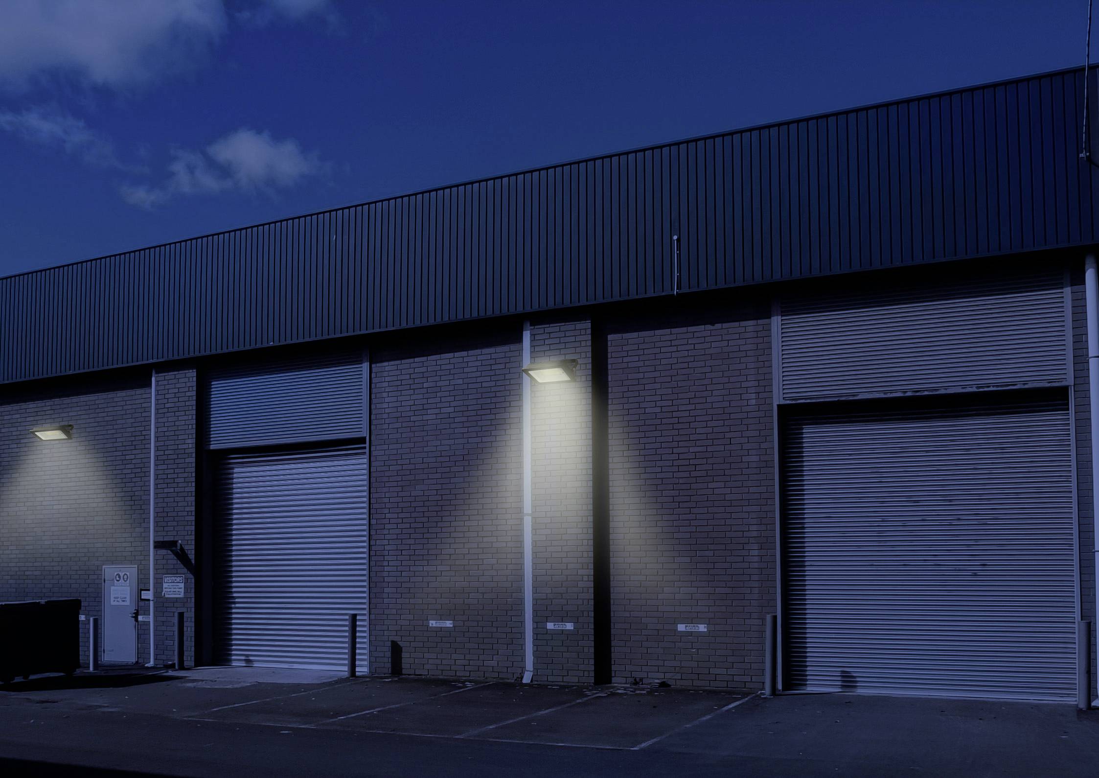 Industriegebäude bei Nacht mit beleuchteten Garagenrolltoren, Betonwänden und teils wolkenverhangenem Himmel im Hintergrund.