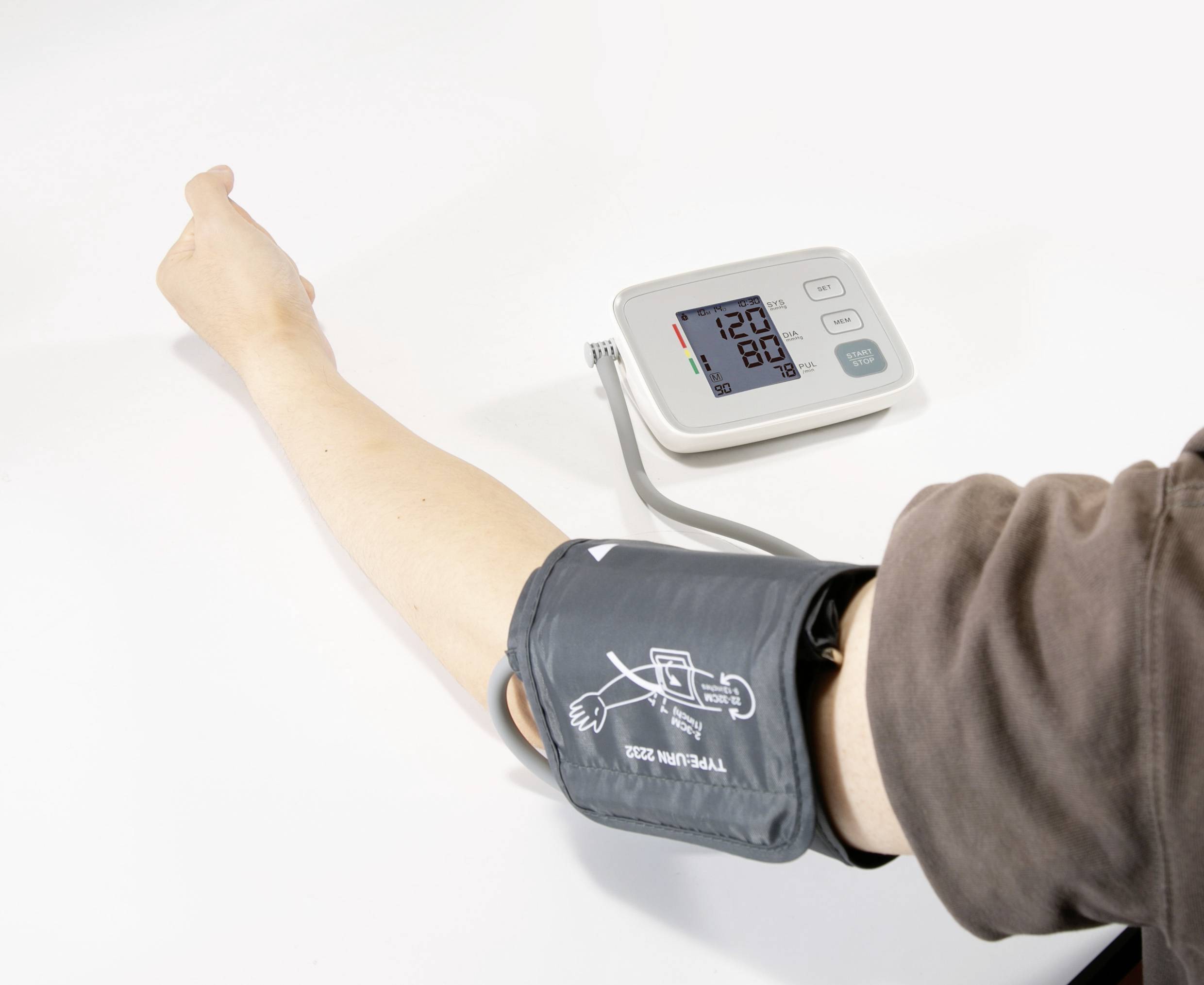 Eine Person misst den Blutdruck mit einem digitalen Gerät. Der Bildschirm zeigt 120 zu 80. Ein Arm liegt ausgestreckt auf einem Tisch.