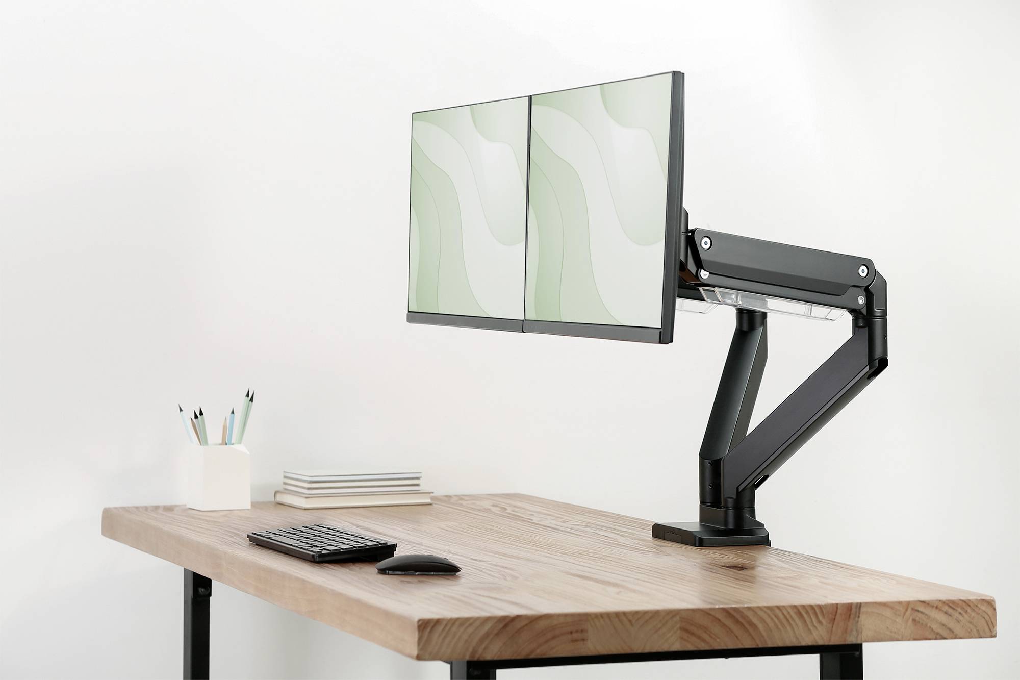 Ein Holzschreibtisch mit zwei Monitoren auf einem Gelenkarm, einer Tastatur, einer Maus und einem Stifthalter mit Stiften; klare, minimalistische Einrichtung.