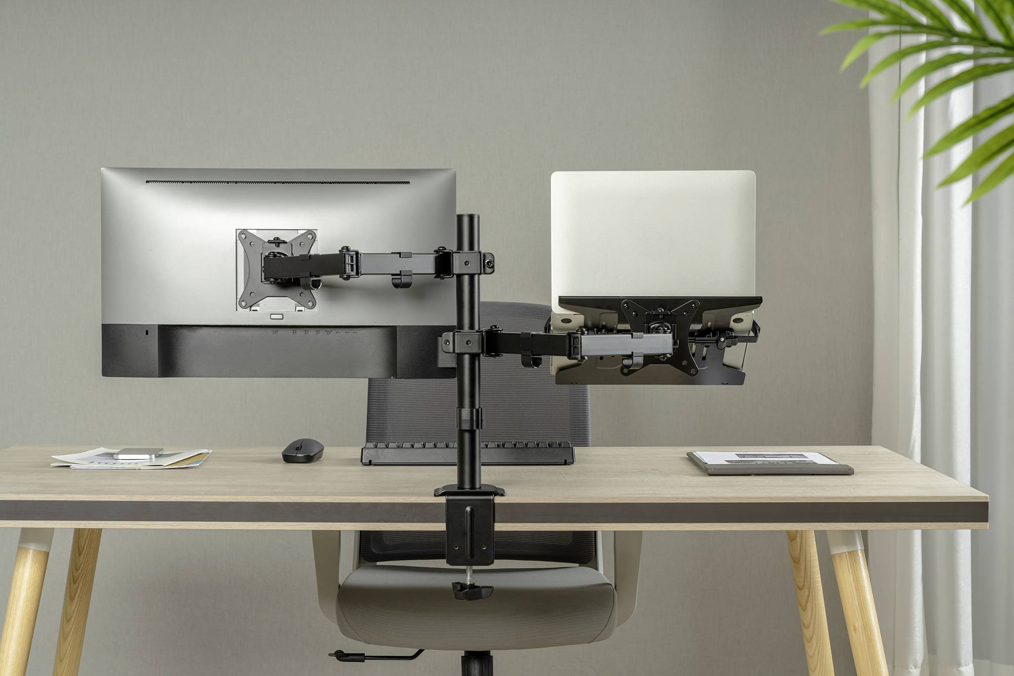Zwei Monitore an einem Doppelarm-Halter auf einem Schreibtisch, einer zeigt einen Laptop, daneben Büromaterial. Im Hintergrund neutrale Wand.