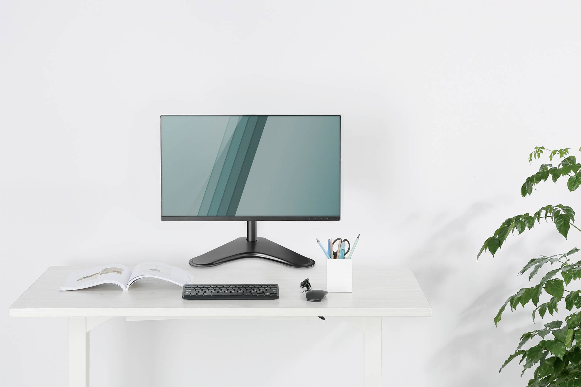 Ein moderner Computermonitor auf einem weißen Schreibtisch. Daneben stehen Tastatur, offenes Buch, Stiftehalter und eine Pflanze.