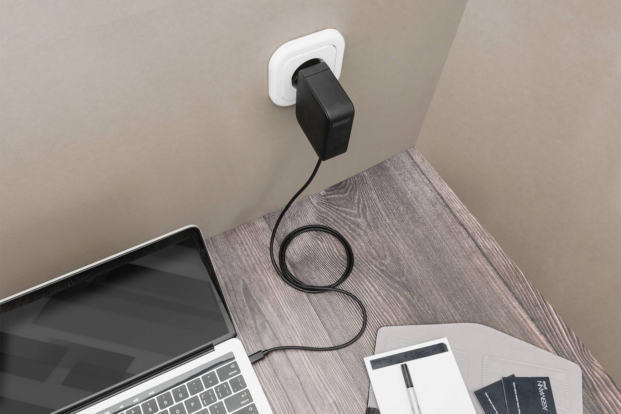 Ein Laptop ist über ein Kabel an eine Steckdose angeschlossen. Auf dem Tisch liegen ein Notizbuch und ein Stift.