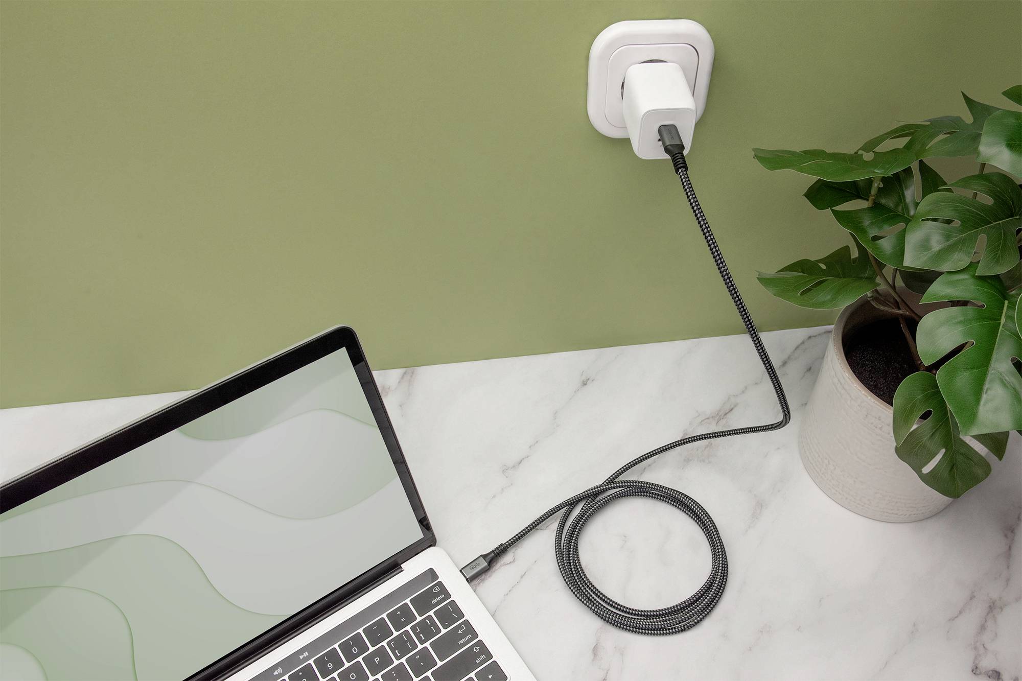 Ein Laptop wird auf einem Marmortisch aufgeladen. Das Ladekabel ist in eine Steckdose an der grünen Wand eingesteckt. Neben dem Laptop steht eine Pflanze.