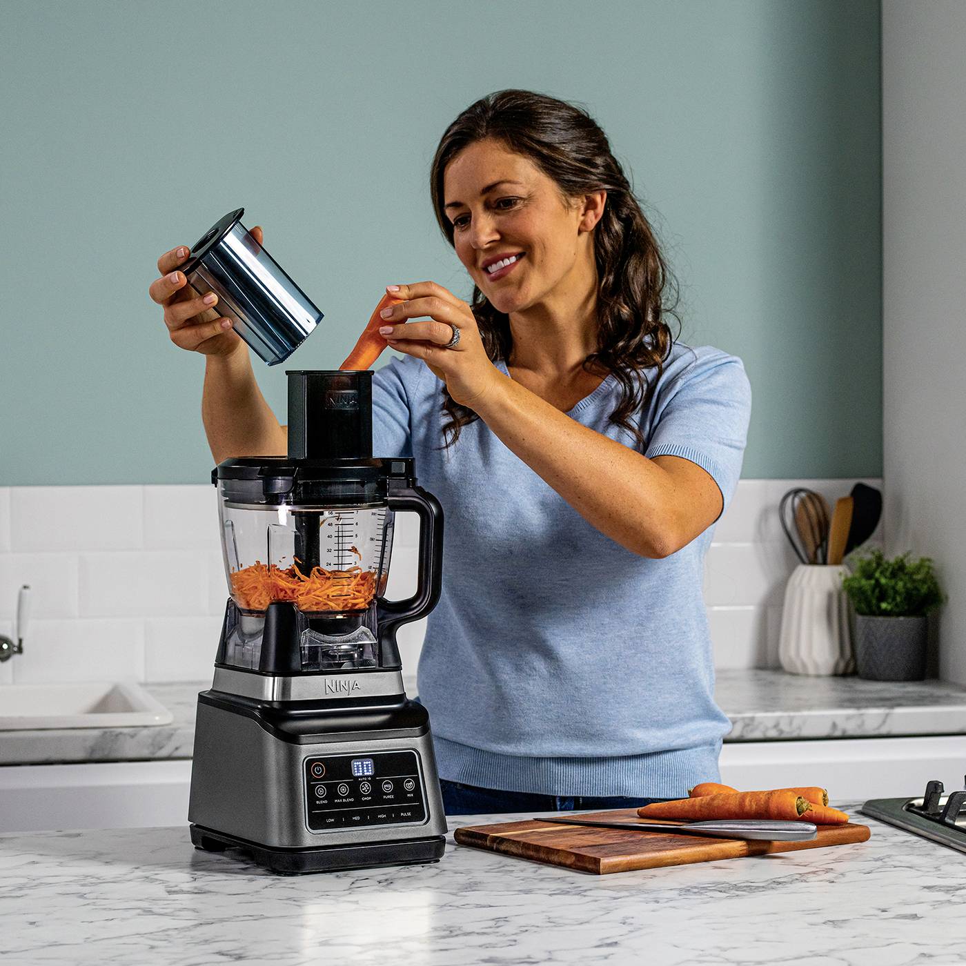 Eine Frau benutzt einen Standmixer in einer Küche. Der Mixer enthält geriebene Karotten. Auf der Arbeitsplatte liegt ein Schneidebrett mit Karotten.