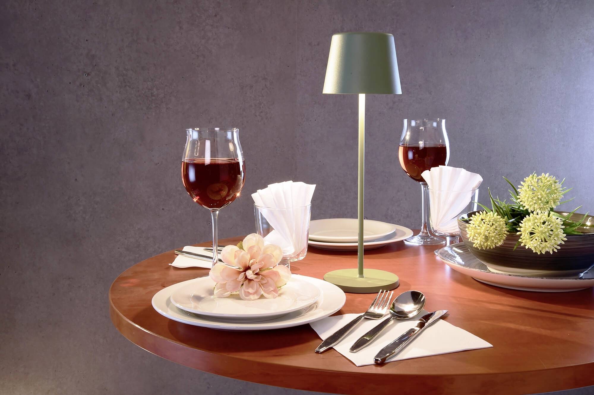Ein runder Tisch ist elegant gedeckt mit zwei Weingläsern, einer blühenden Blume auf einem Teller und einer modernen Tischlampe.