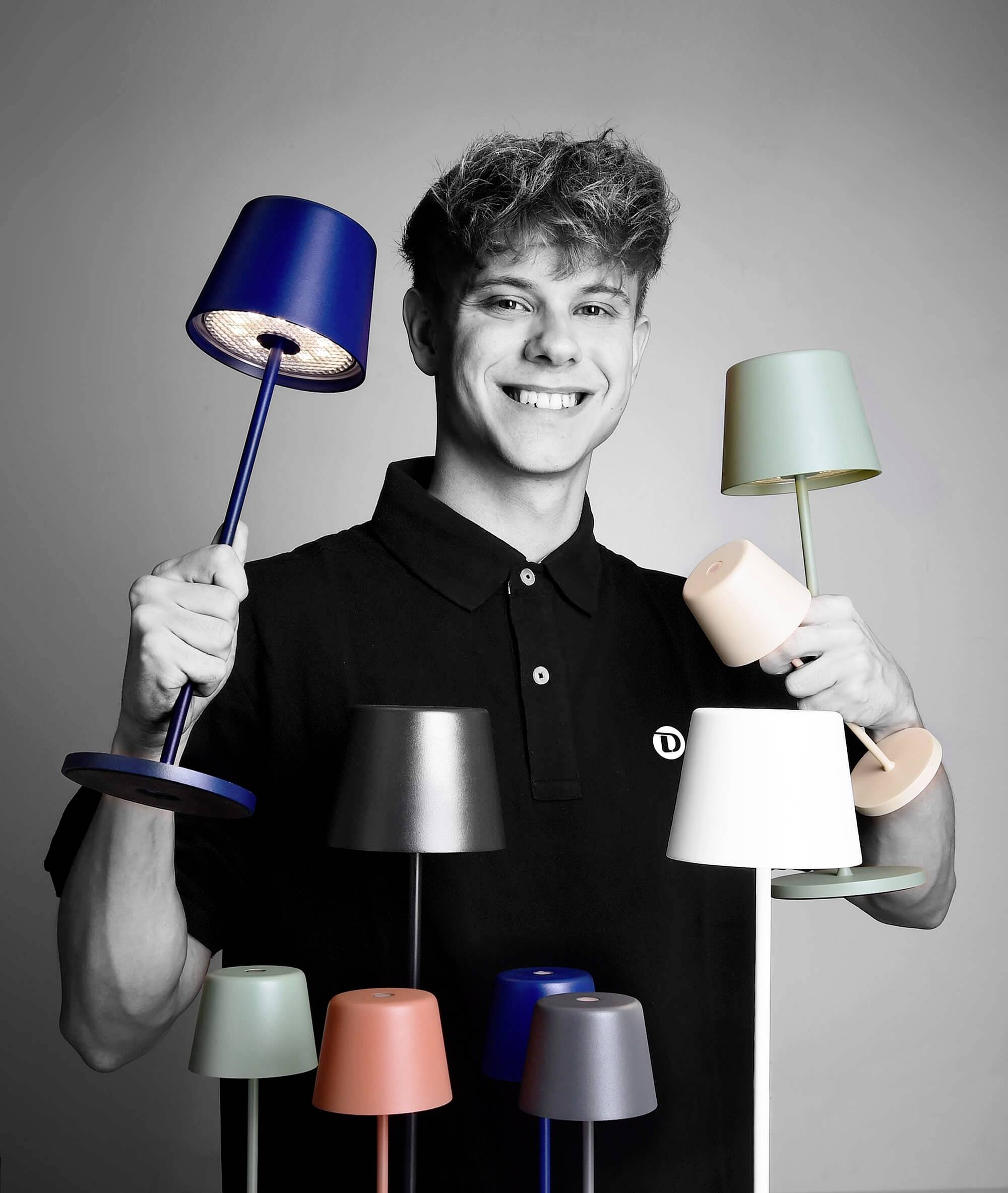 Junger Mann lächelt und hält mehrere bunte Tischlampen in den Händen. Der Hintergrund ist neutral, Fokus auf die Lampen.