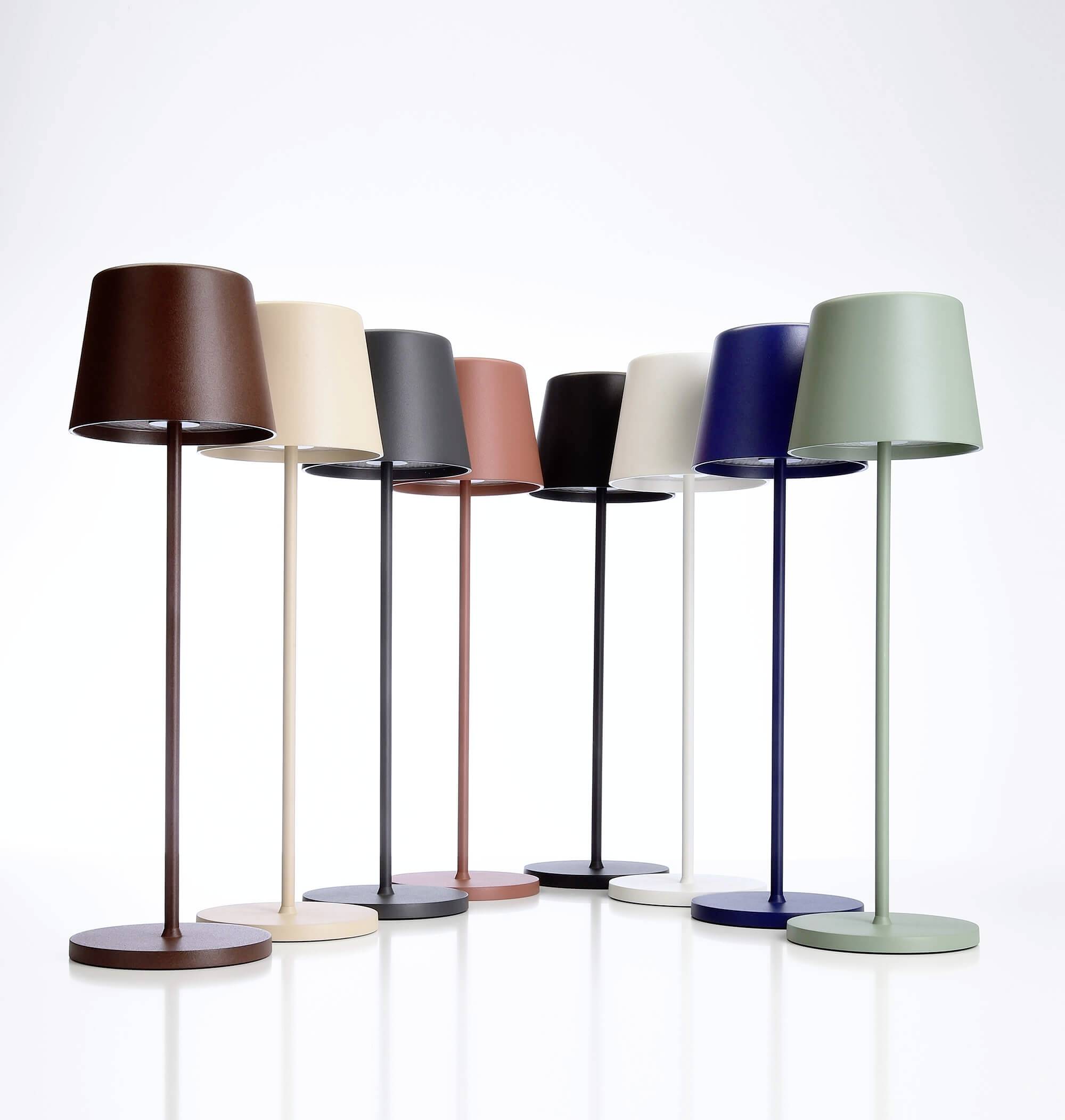 Acht moderne Tischlampen in verschiedenen Farben, aufgereiht vor weißem Hintergrund. Farben umfassen Braun, Beige, Schwarz, Rosa, Blau und Grün.