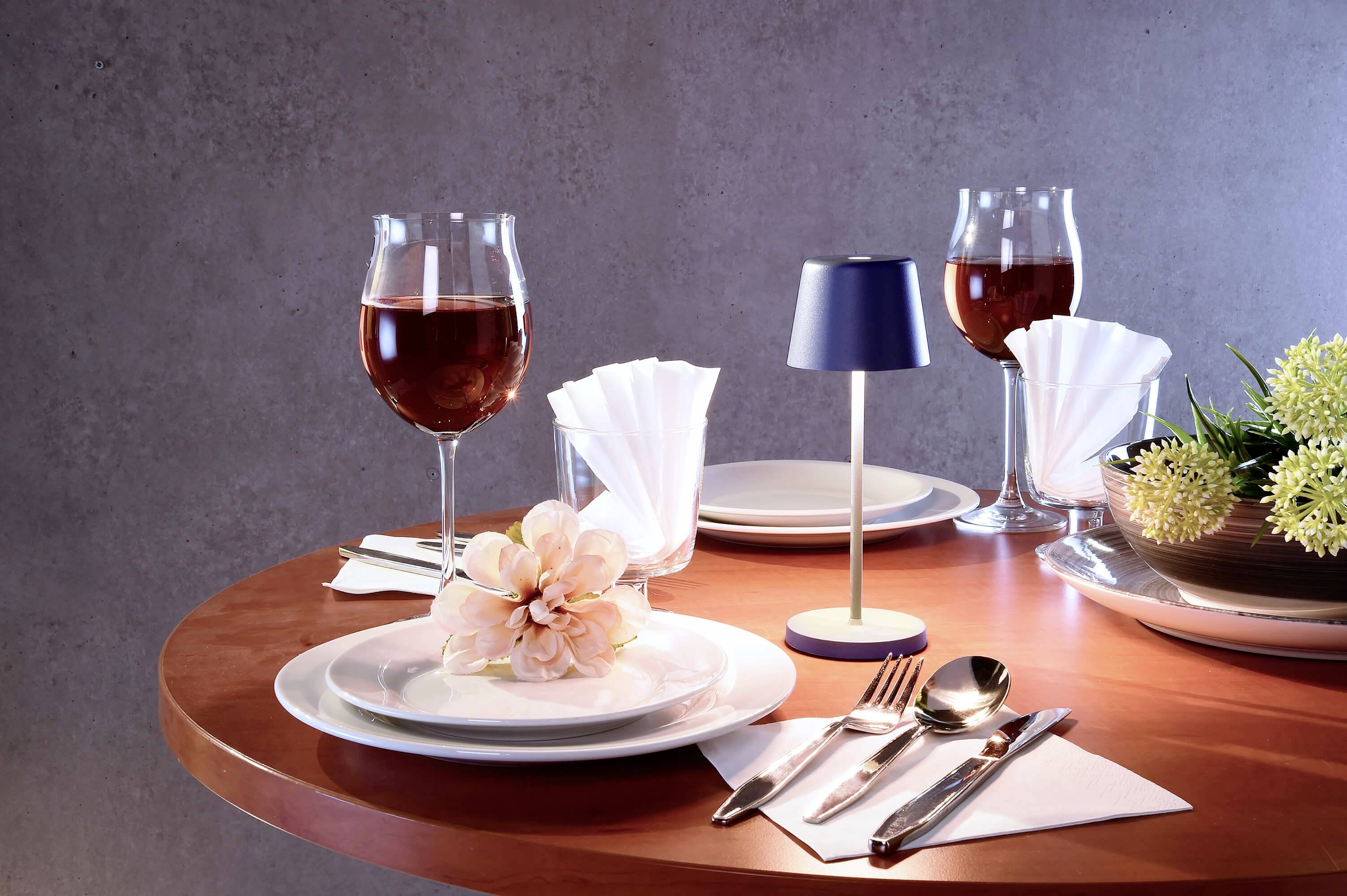 Ein runder Tisch mit zwei gedeckten Plätzen, Weingläsern mit rotem Getränk, gefalteten Servietten, Besteck, Blumen und Tischlampe.