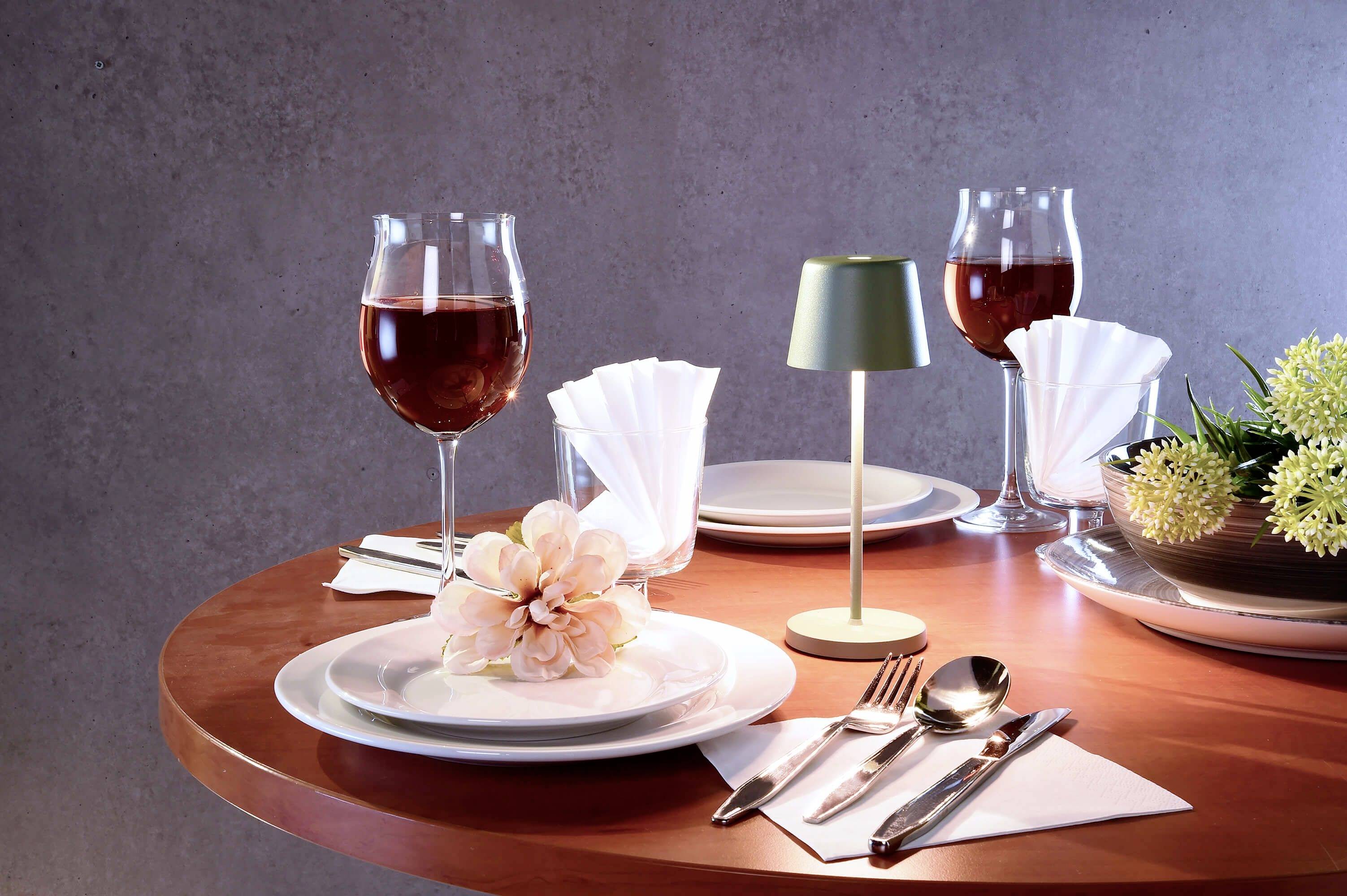 Ein gedeckter Tisch mit Weingläsern, Tellern, Besteck und einer kleinen Tischlampe. Dekorativ arrangierte Blumen und Papierservietten.