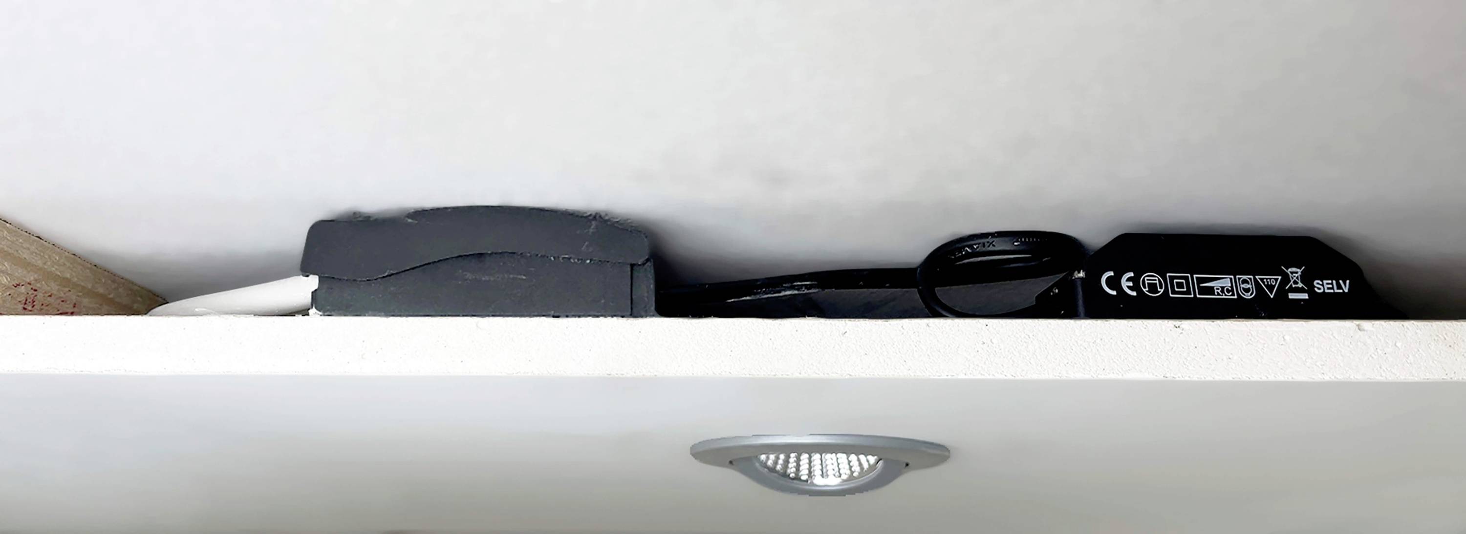 Ein schwarzes Elektronikgerät mit Kabeln auf einem weißen Regal, darüber sind verschiedene Symbole und Schriftzüge erkennbar.