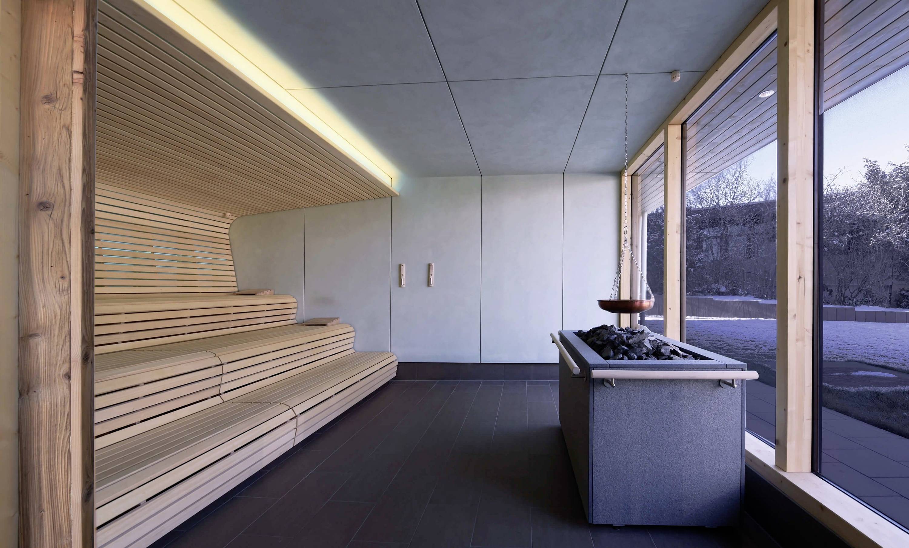 Moderne Sauna mit Holzverkleidung, Sitzbänken und einem Saunaofen. Große Fenster geben Blick nach draußen auf verschneite Landschaft.