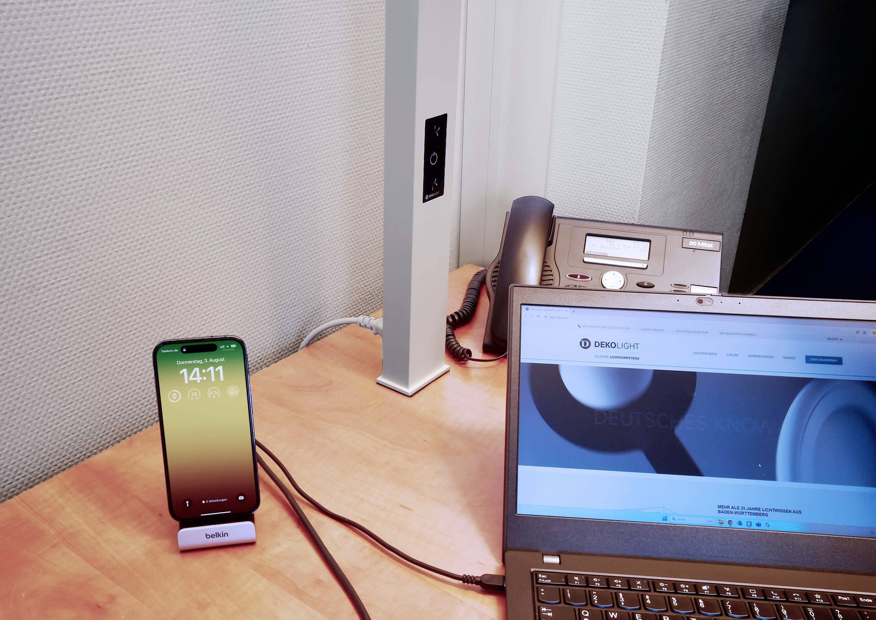 Ein Schreibtisch mit einem Laptop, einem Smartphone auf einem Ladegerät und einem Telefon. Der Laptop zeigt eine Website an.