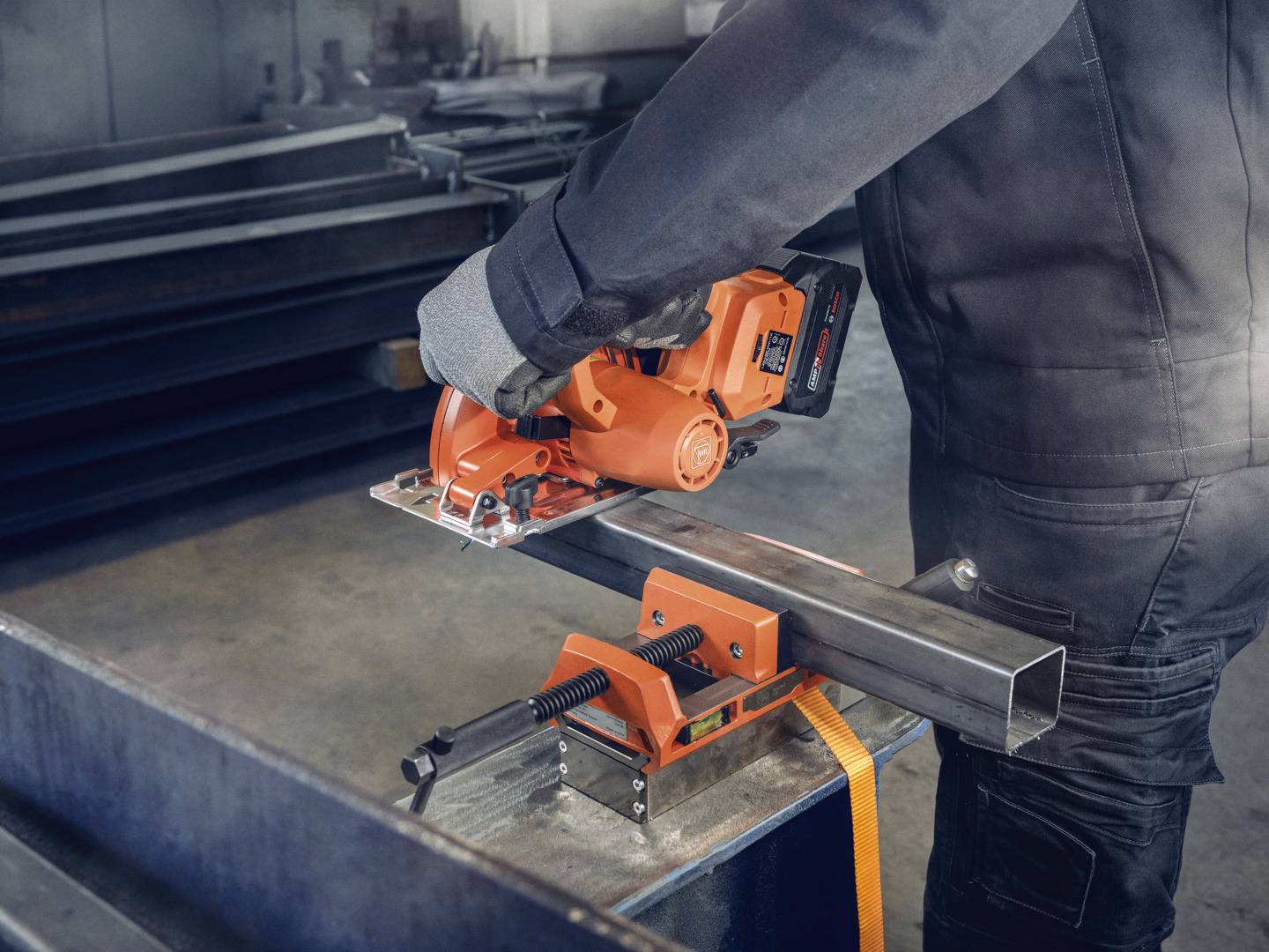 Person arbeitet mit einer orangefarbenen, tragbaren Metallsäge, um ein quadratisches Metallrohr zu schneiden. Schutzkleidung wird getragen.