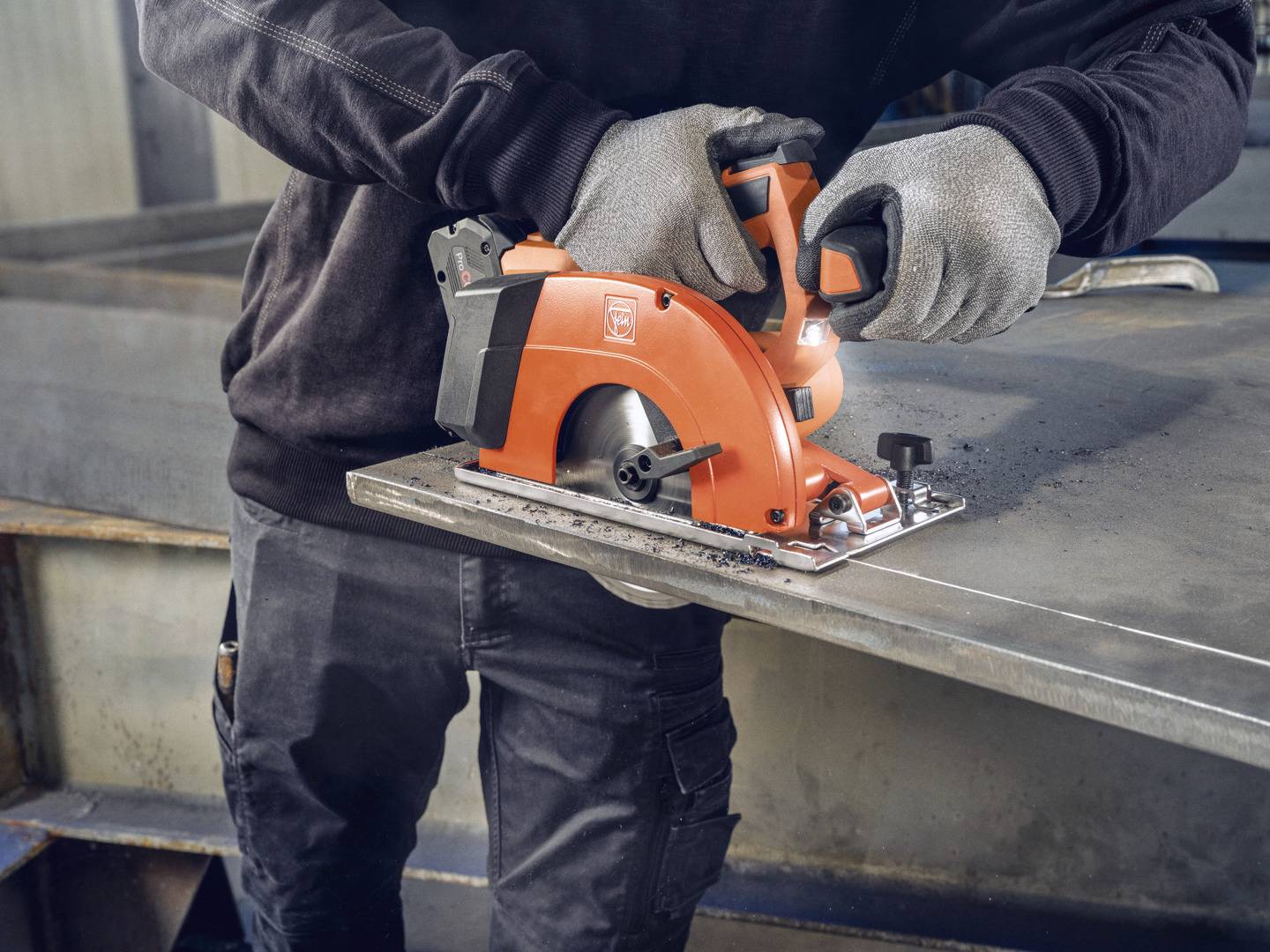 Person sägt mit einer orangefarbenen Kreissäge Metallplatte. Schutzkleidung wird getragen. Prozess der Bearbeitung von Metall sichtbar.
