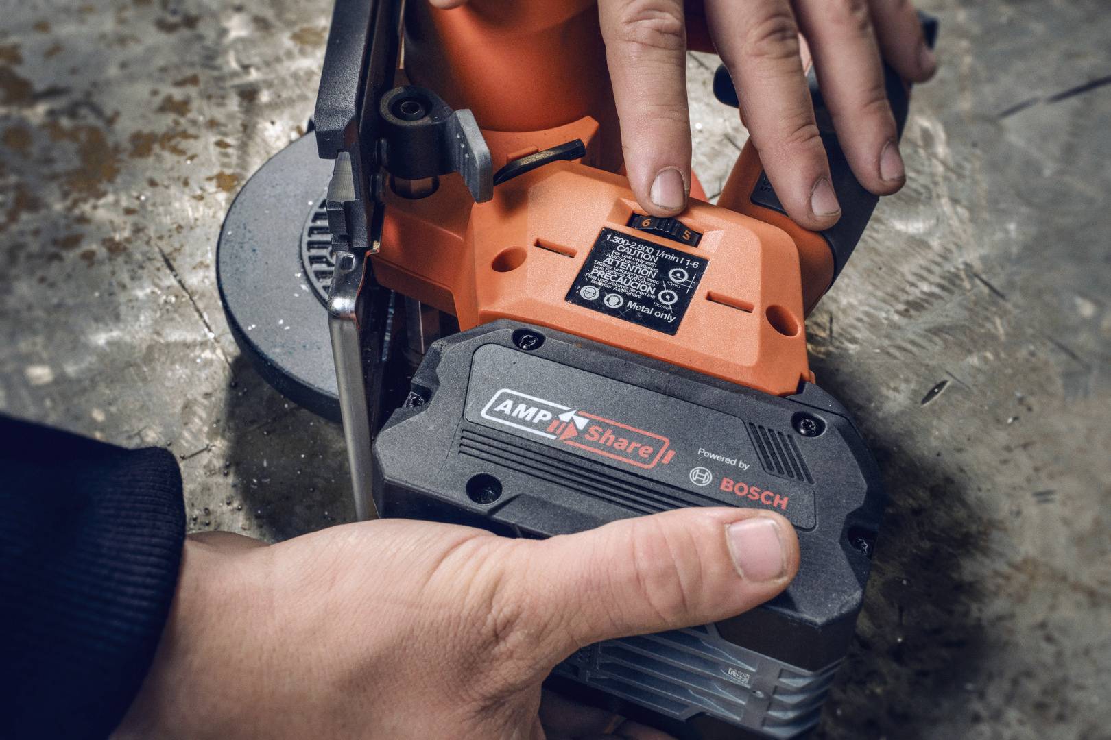Eine Person hält ein elektrisches Werkzeug mit einem 'AmpShare'-Akku von Bosch. Die Hände justieren das Gerät auf einer Metalloberfläche.