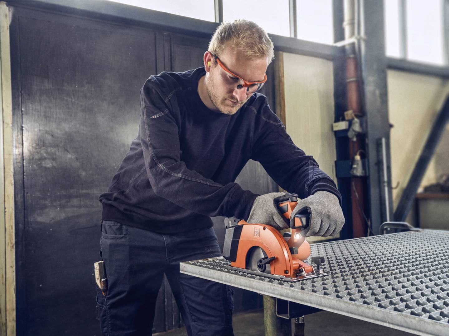 Eine Person trägt Schutzbrille und Handschuhe, während sie mit einer Kreissäge Metall auf einer Werkbank in einer Werkstatt schneidet.