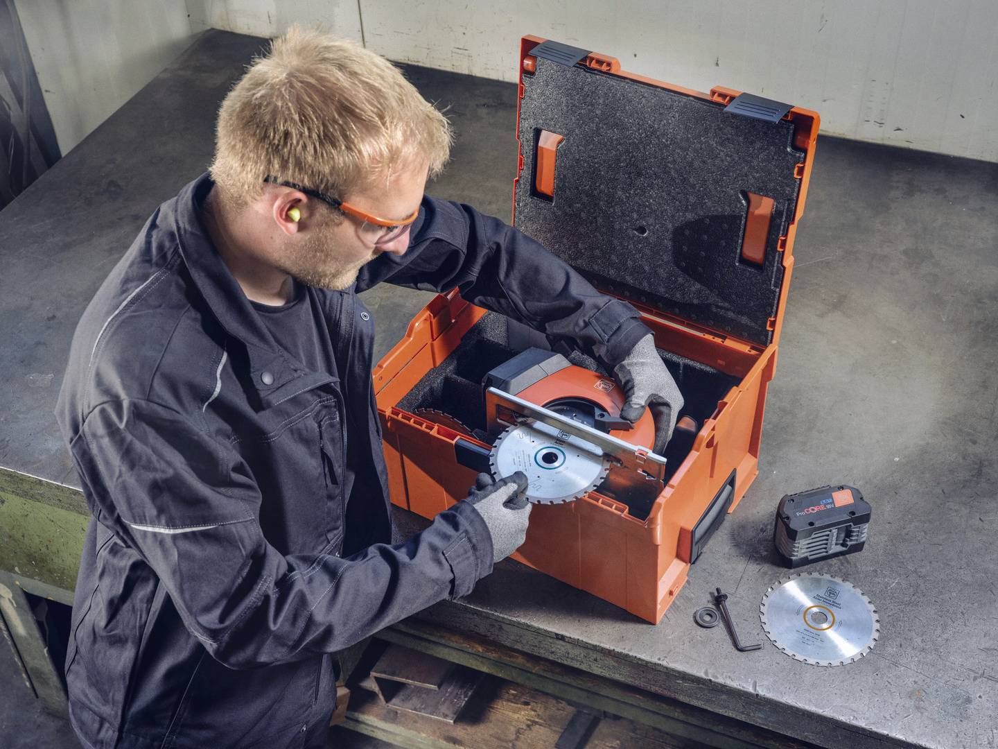 Ein Mann in Arbeitskleidung wechselt die Schleifscheibe einer Maschine. Auf dem Tisch befinden sich ein Ersatzblatt und eine Batterie.